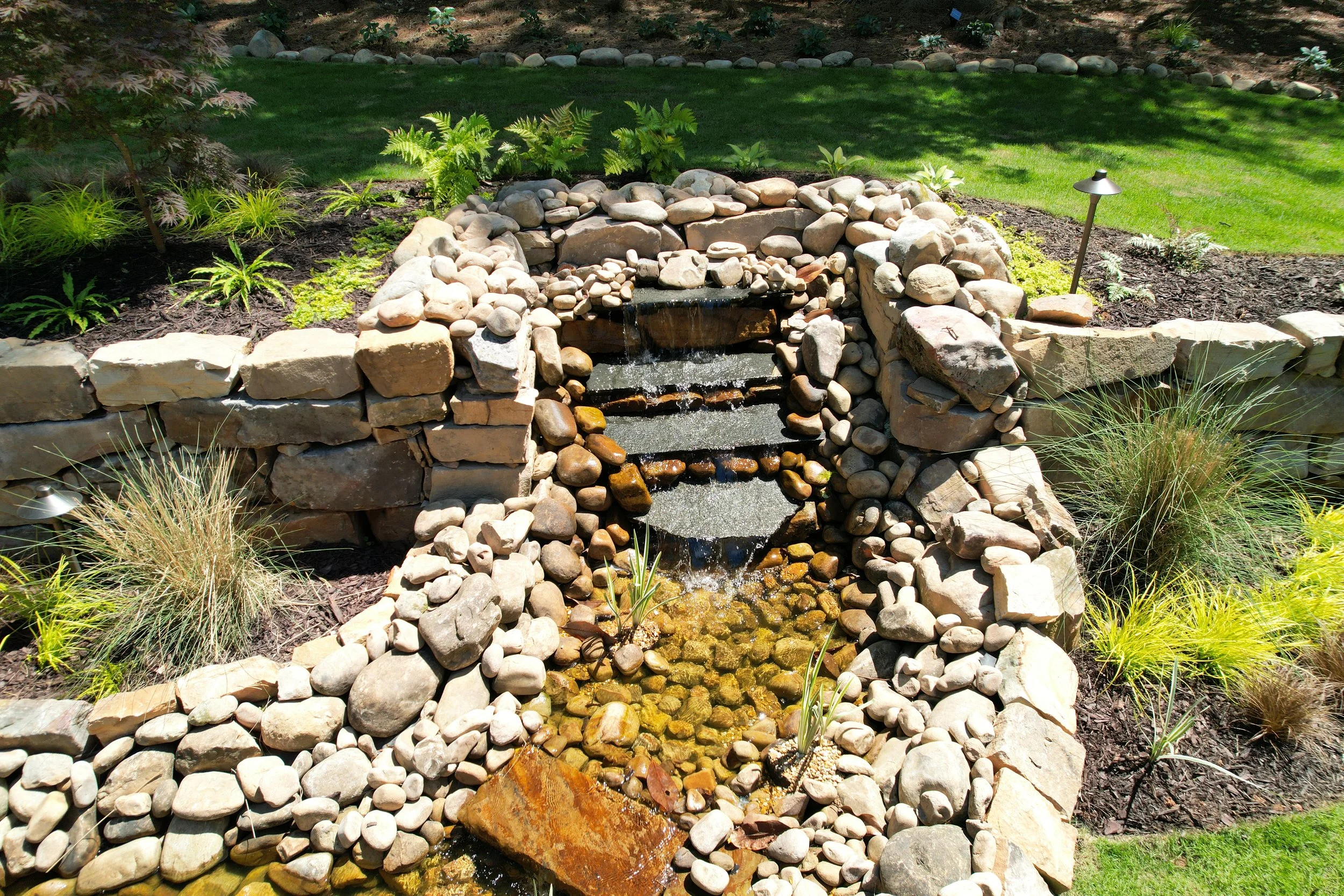 Tiered stone and river rock waterfall feature with cascading water steps and landscape lighting.