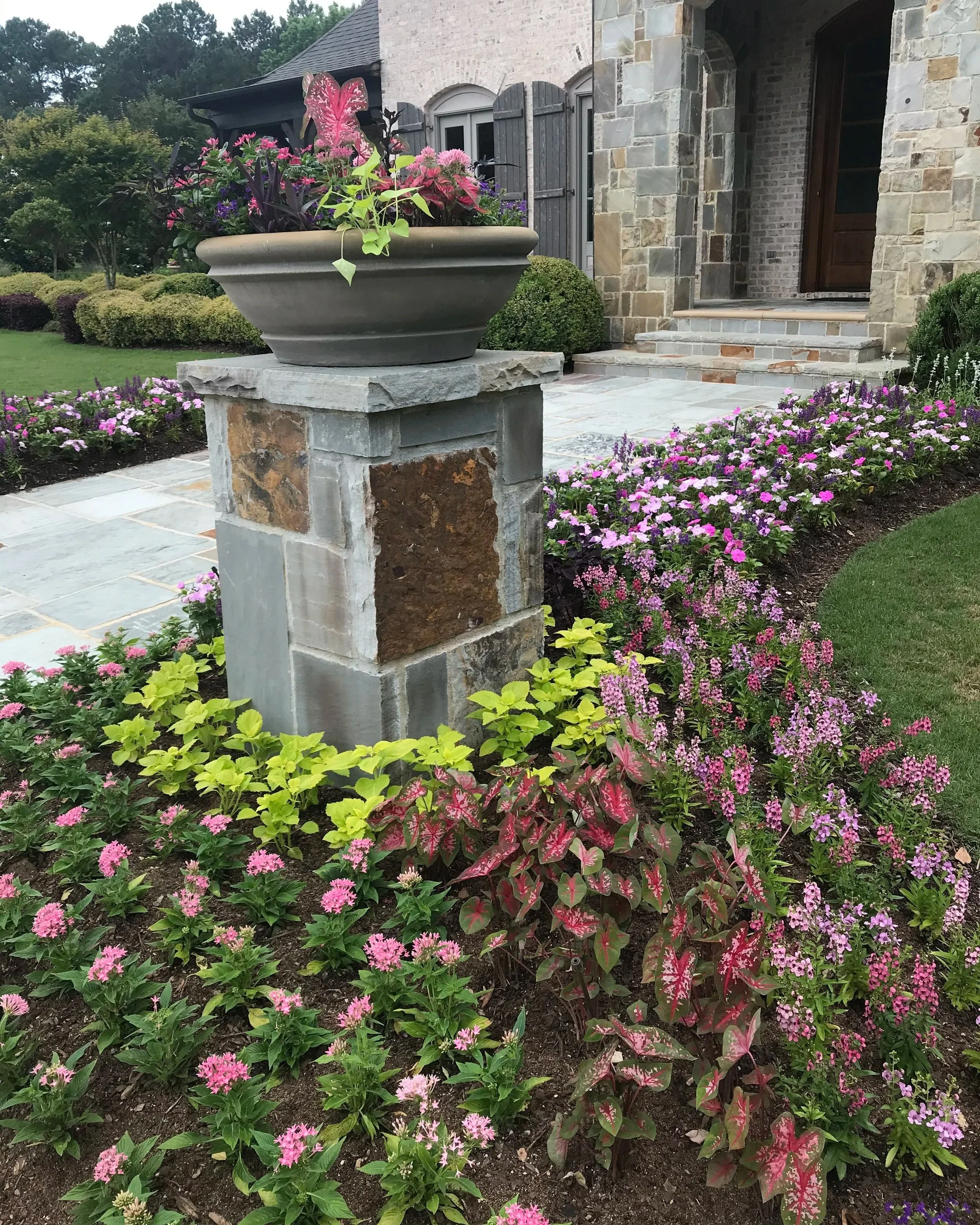 Professionally installed front entry landscape bed with colorful annuals, caladiums, and a decorative stone planter column at a stone home in Athens Georgia by Southern Grounds Landscaping
