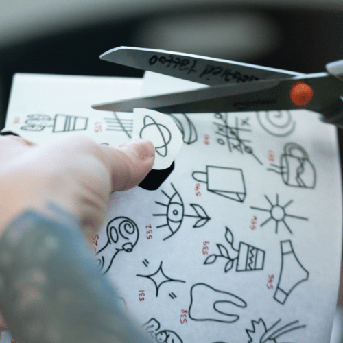 hand holding a sheet of pre-drawn tattoo flash designs and cutting one out with scissors to prepare for the tattoo
