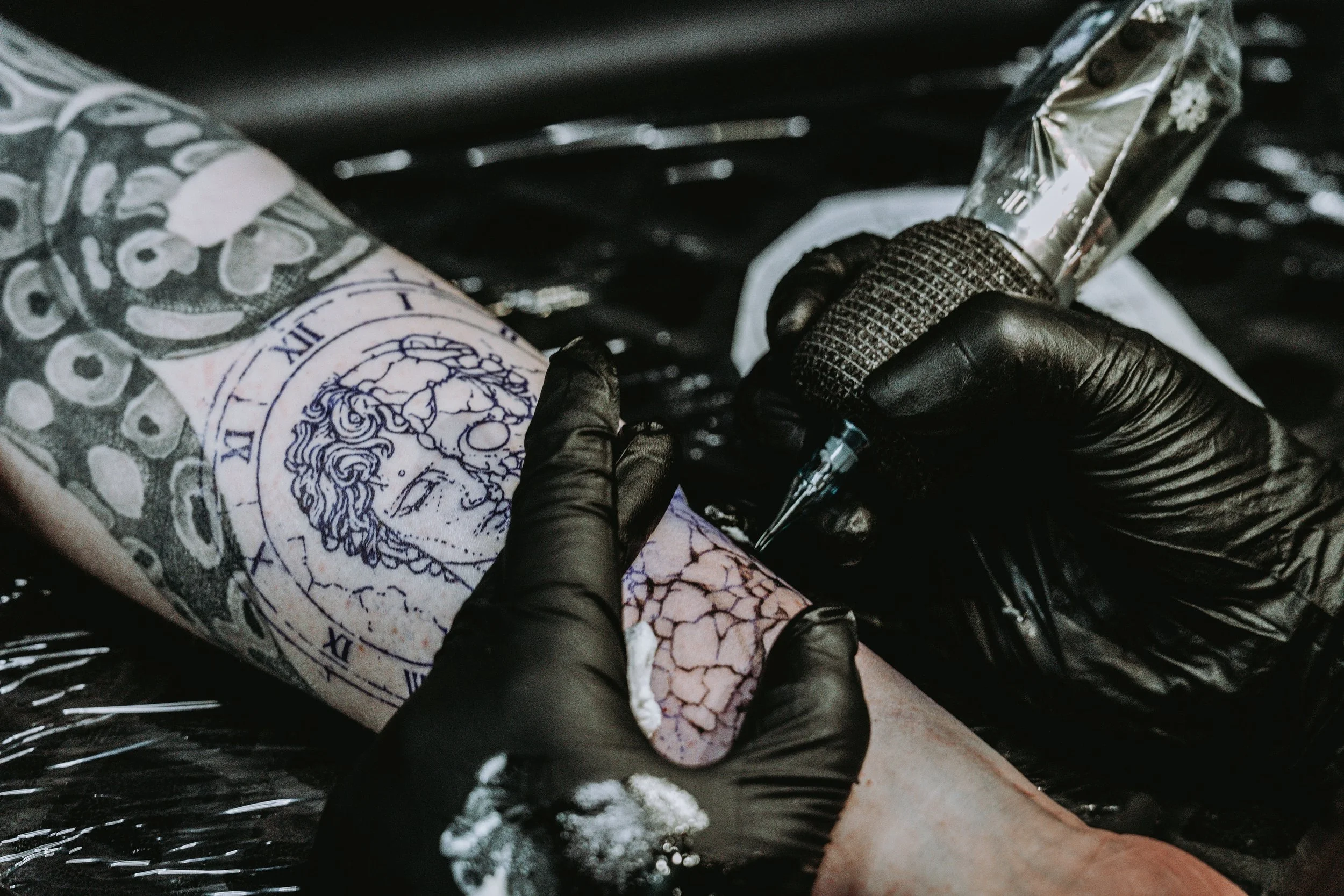 hands in black gloves holding a tattoo machine doing a tattoo on a persons forearm