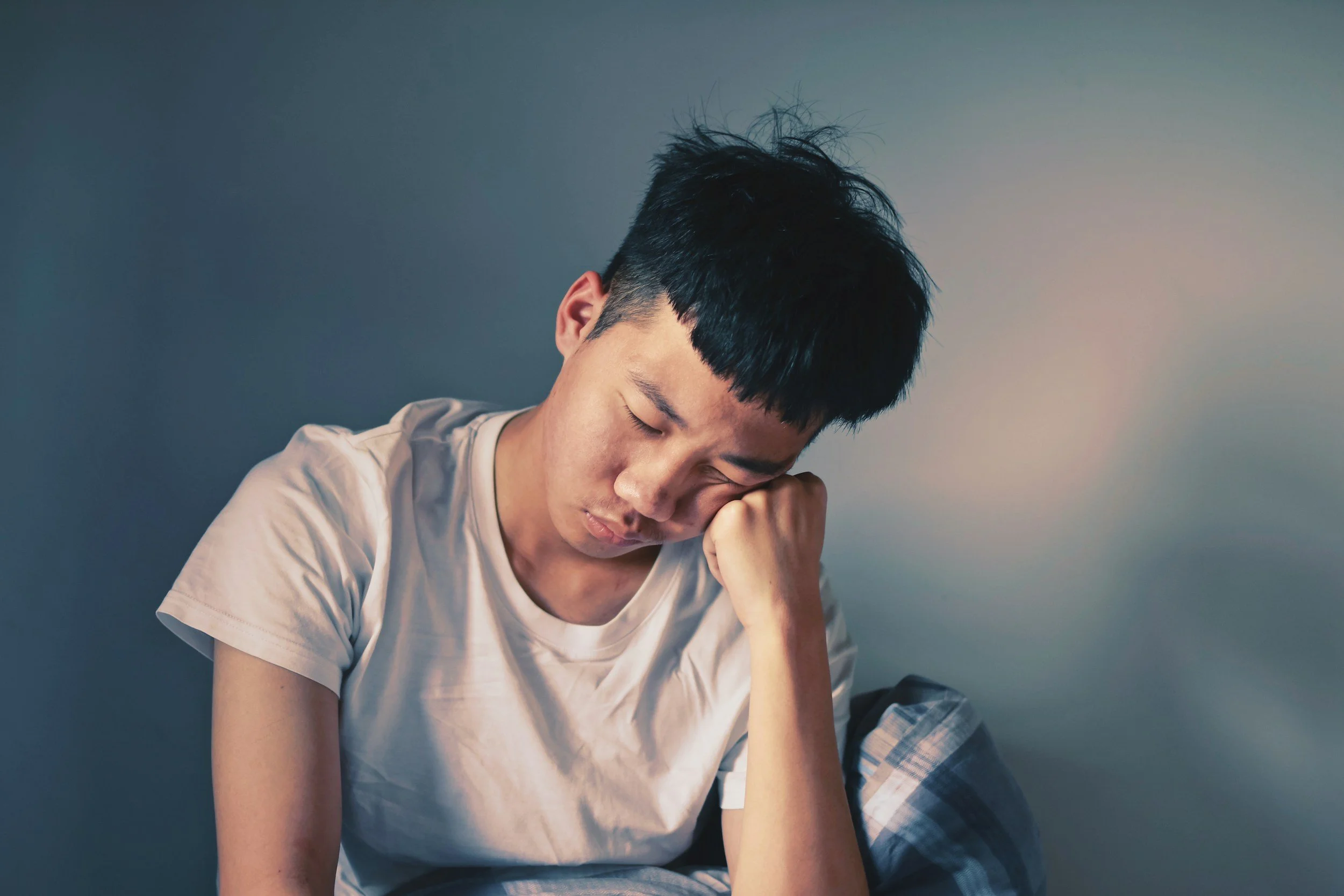 Asian male in pajamas looking down resting faceon his fist appearing having difficulty coping with stress