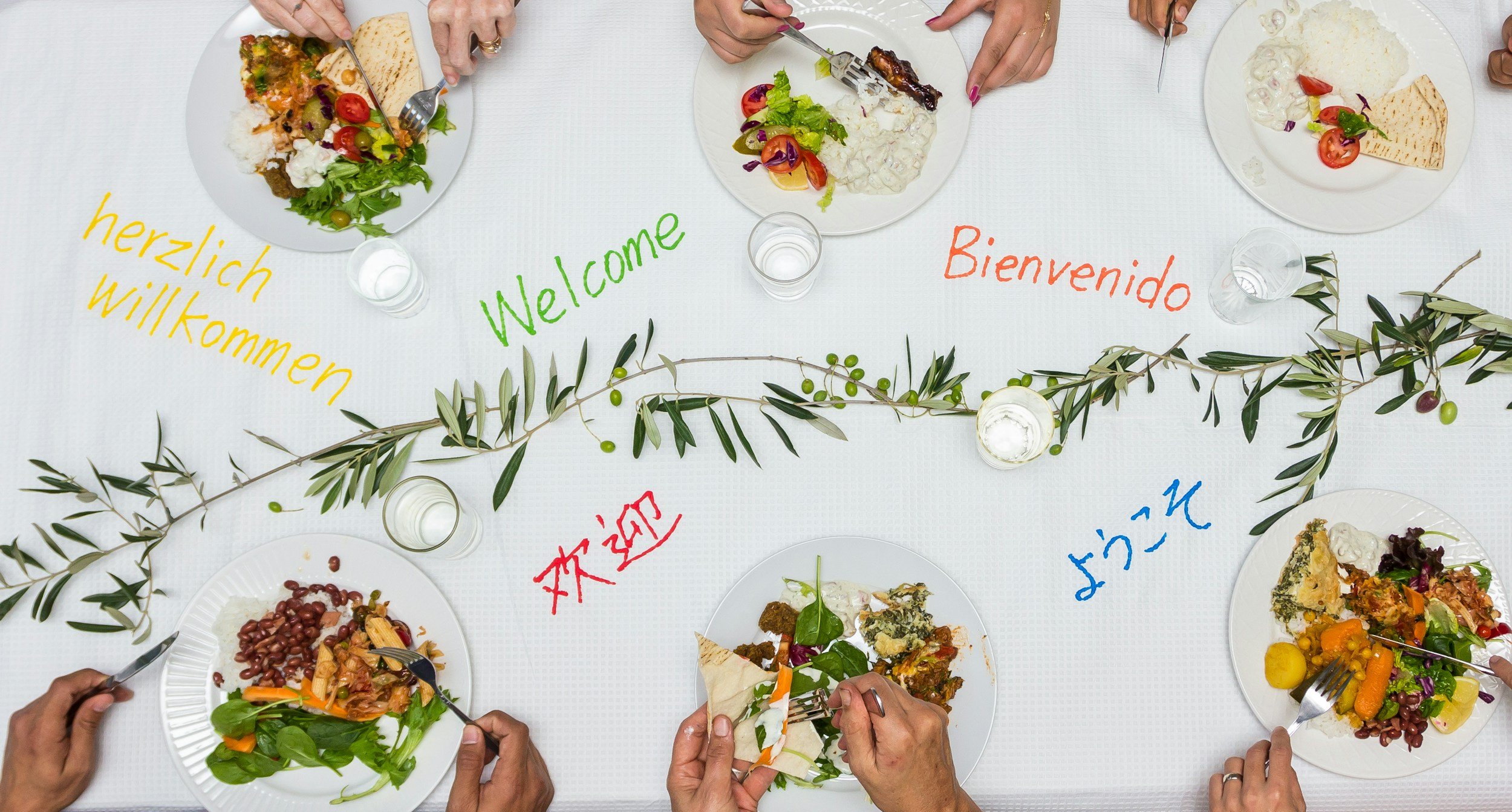 plates of food and hands on table cloth that has the word "welcome" in different languages