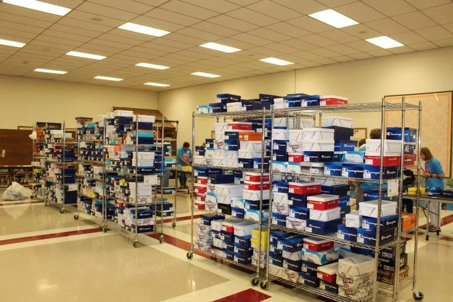 Large room with metal shelves filled with many shoeboxes, people working at tables in the background, ceiling with fluorescent lights.