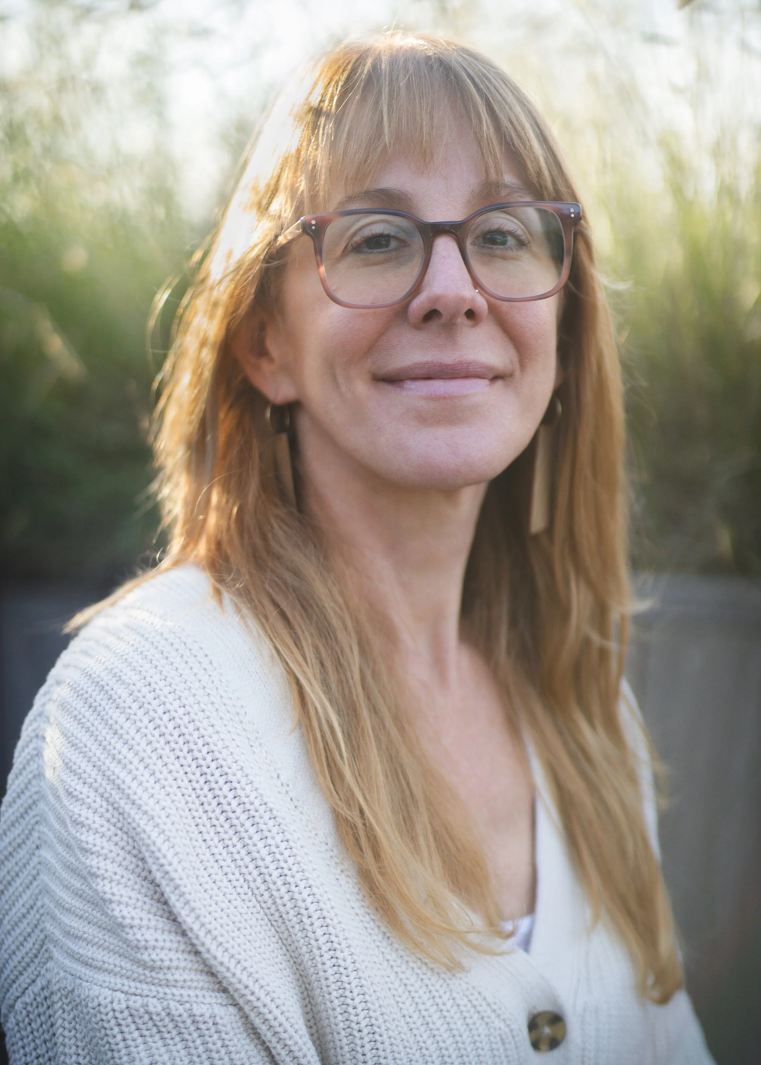 A woman with blonde hair, wearing glasses and a gray knit hat, smiling and looking at a small dog dressed in a white-colored jacket, sitting with tall grass in the background