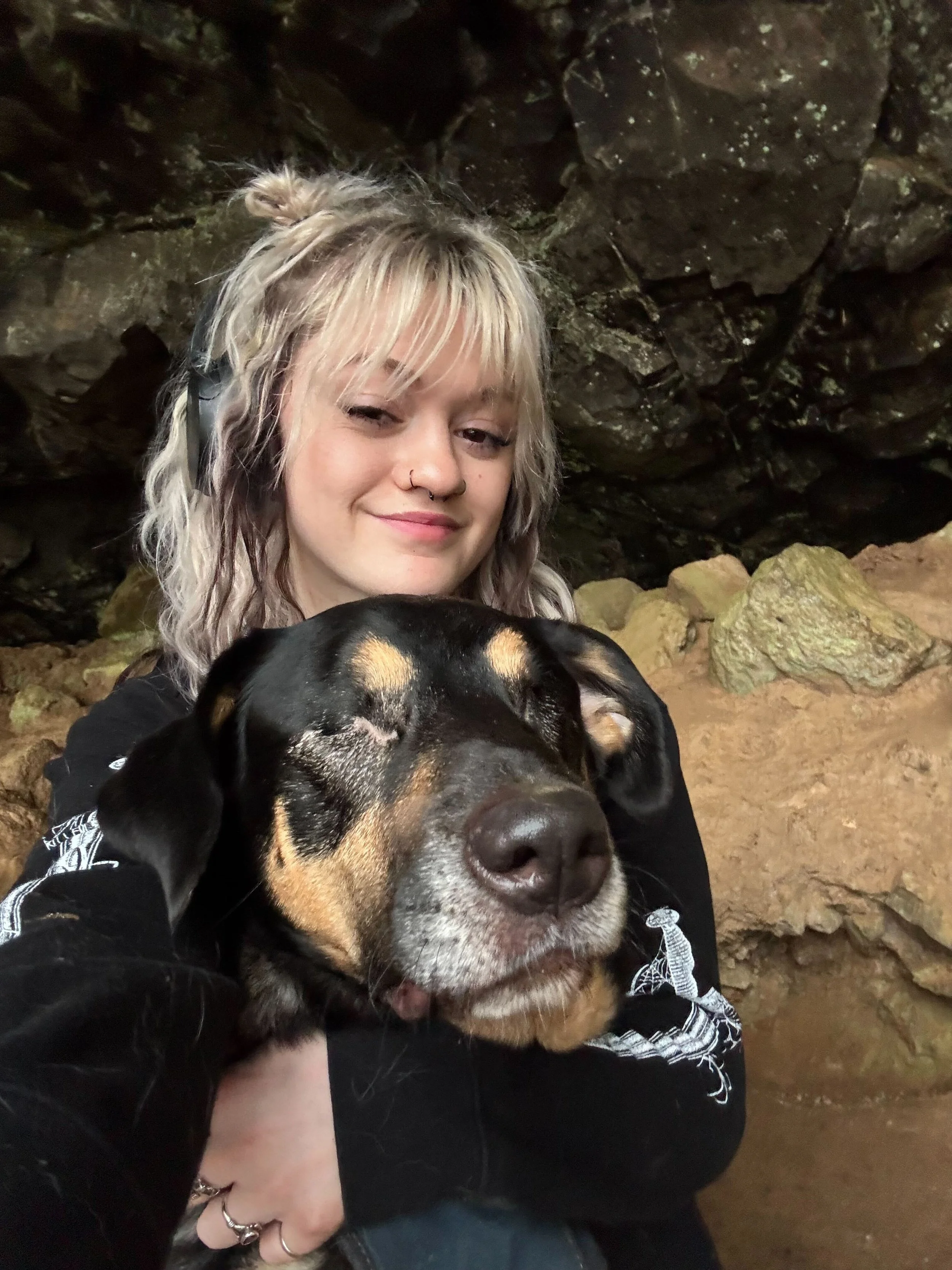 A young woman with blonde hair with black headphones, sitting on rocks, hugging a happy black and tan dog while taking a selfie.