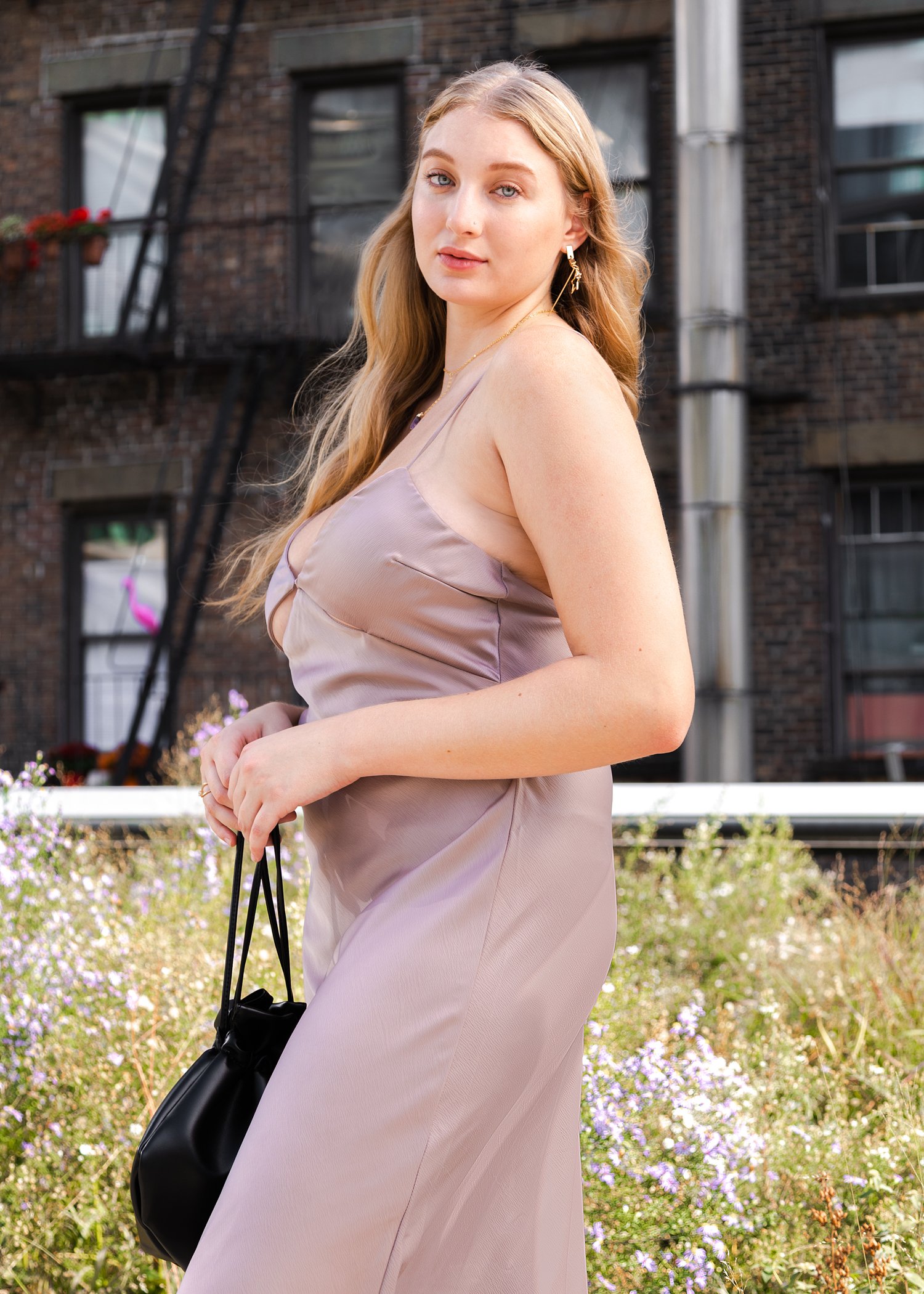 A young woman with long blonde hair wearing a silky taupe slip dress, holding a small black handbag, standing outdoors in front of a brick building and flowering plants.