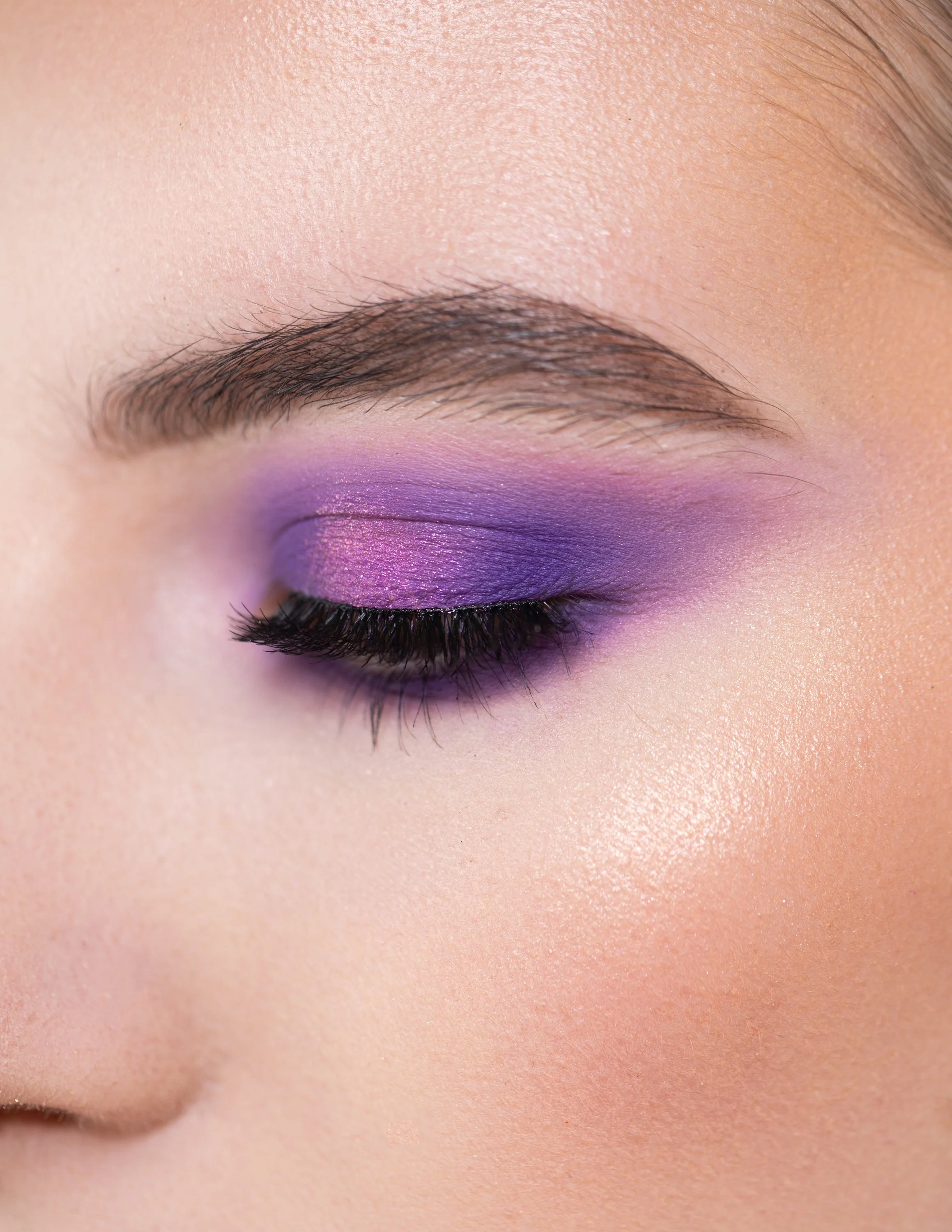 Close-up of a woman's eye with purple eyeshadow and long eyelashes, with smooth skin and well-groomed eyebrow.