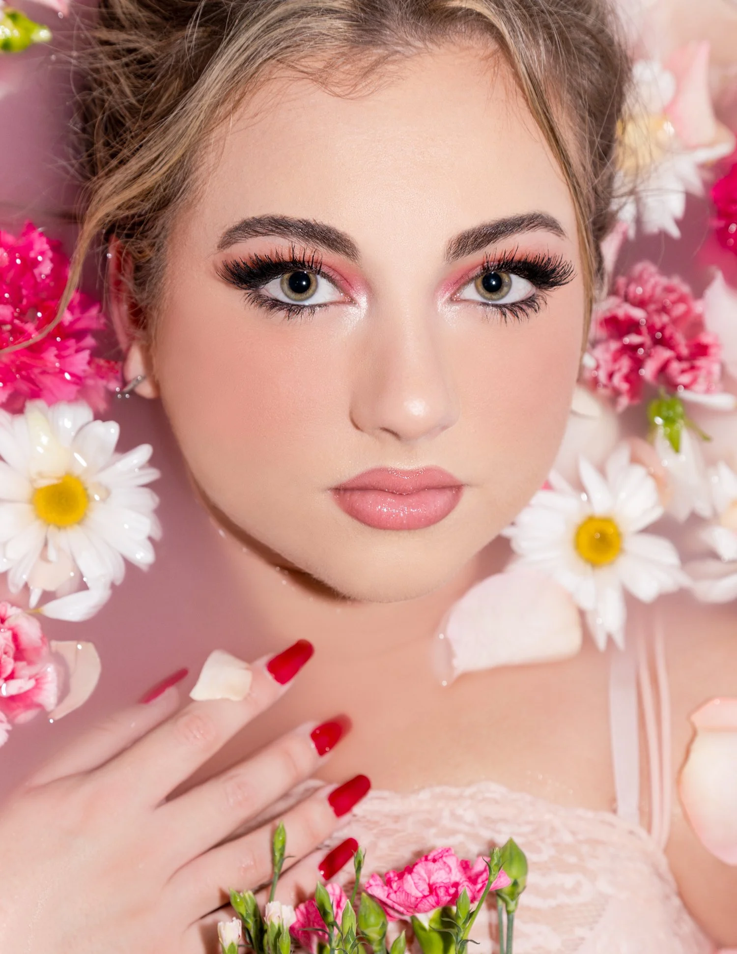 A woman with makeup and red nails surrounded by pink and white flowers, reclining in a bath of water.