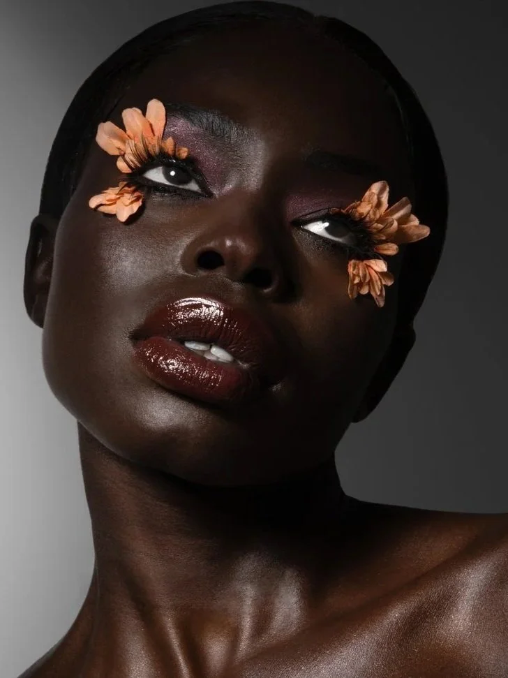 A portrait of a woman with dark skin and short hair, featuring artistic makeup with flower designs around her eyes, glossy lips, and a sleek appearance.