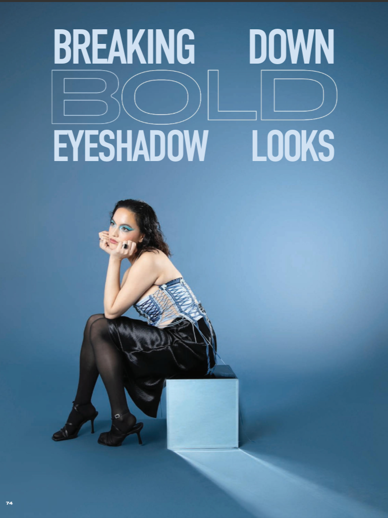 A woman with dark curly hair, sitting on a blue box against a blue background, wearing a stylish strapless top, black skirt, black stockings, and high heels. She has bold blue eyeshadow and is resting her chin on her hand, looking thoughtfully at the