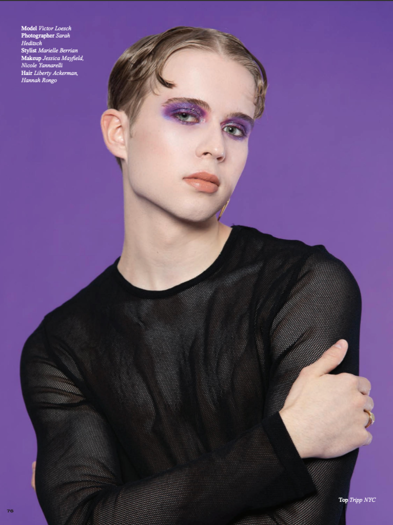 Portrait of a young man with short hair, wearing a black sheer top, against a purple background, with makeup including purple eyeshadow and nude lipstick.