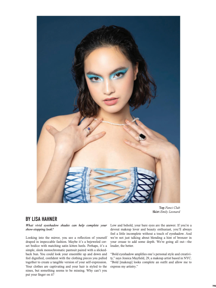 A woman with short dark curly hair and blue eyeshadow posing in front of a blue background. She wears a strapless dress with blue and white laced-up detail, a pearl necklace, and rings on her fingers.