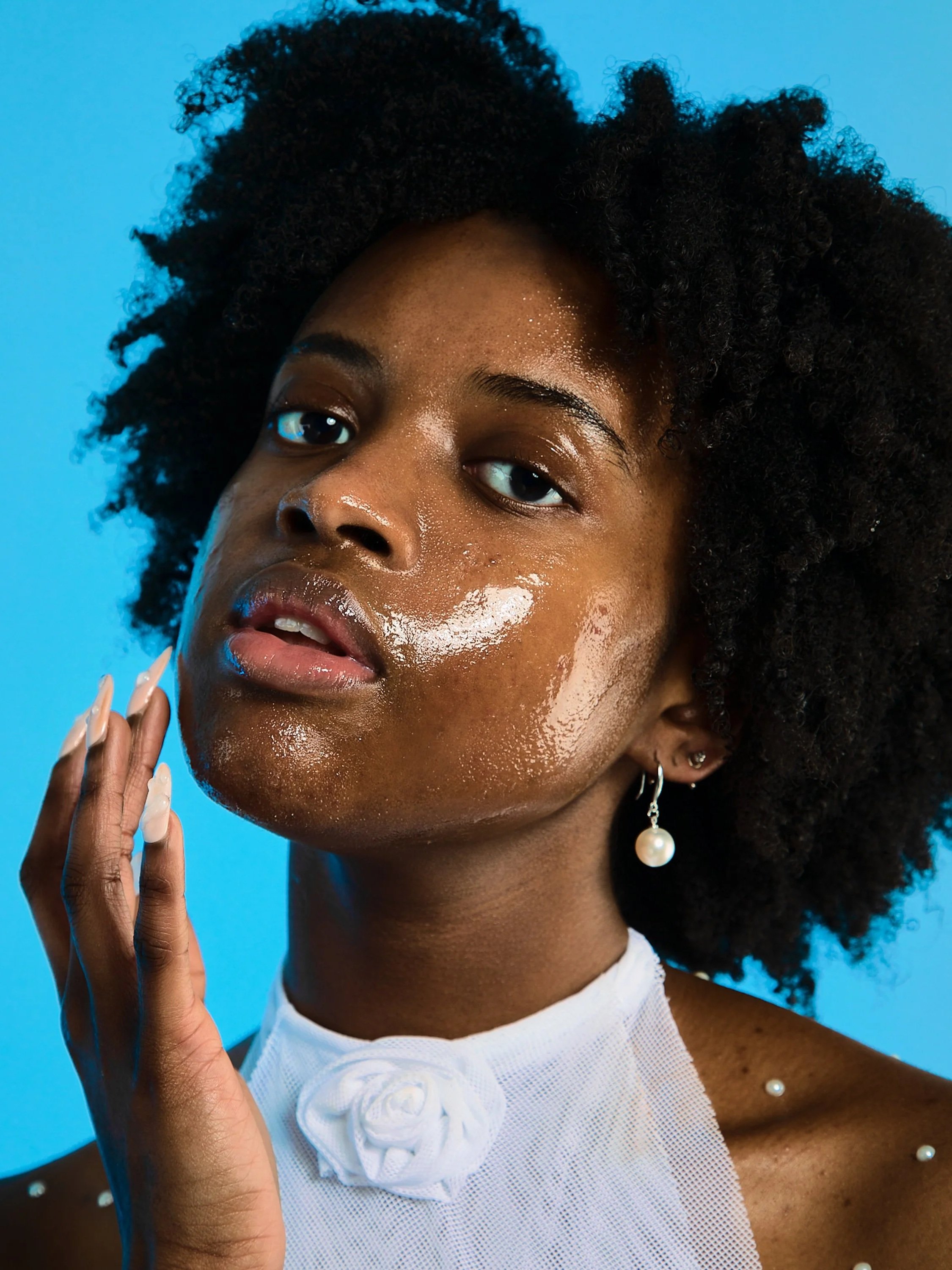 Close-up of a young woman with natural hair, with shiny skin and wearing pearl earrings, a white top with a flower detail, against a blue background.