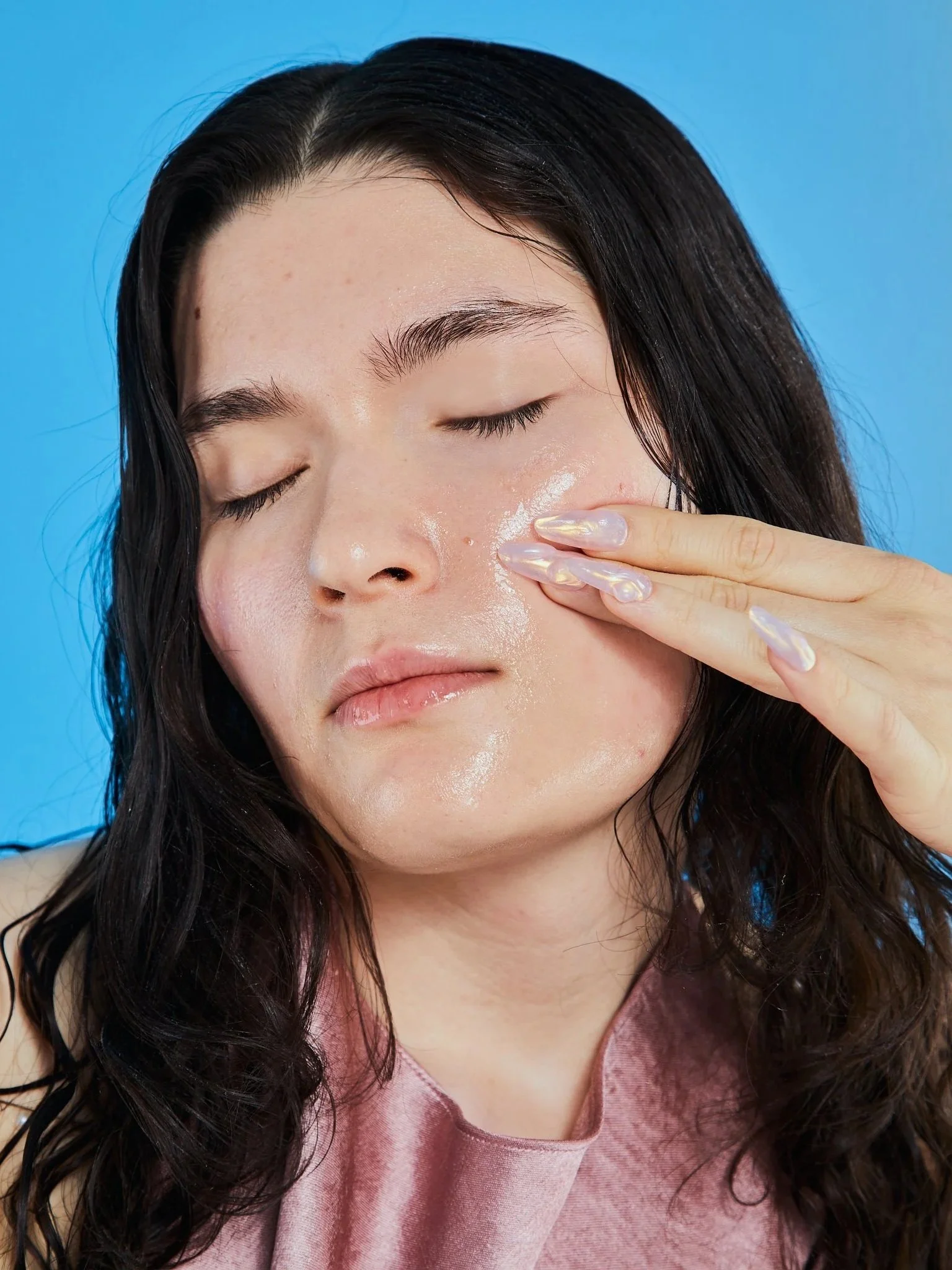 A woman with dark hair applying skincare product to her face against a blue background.