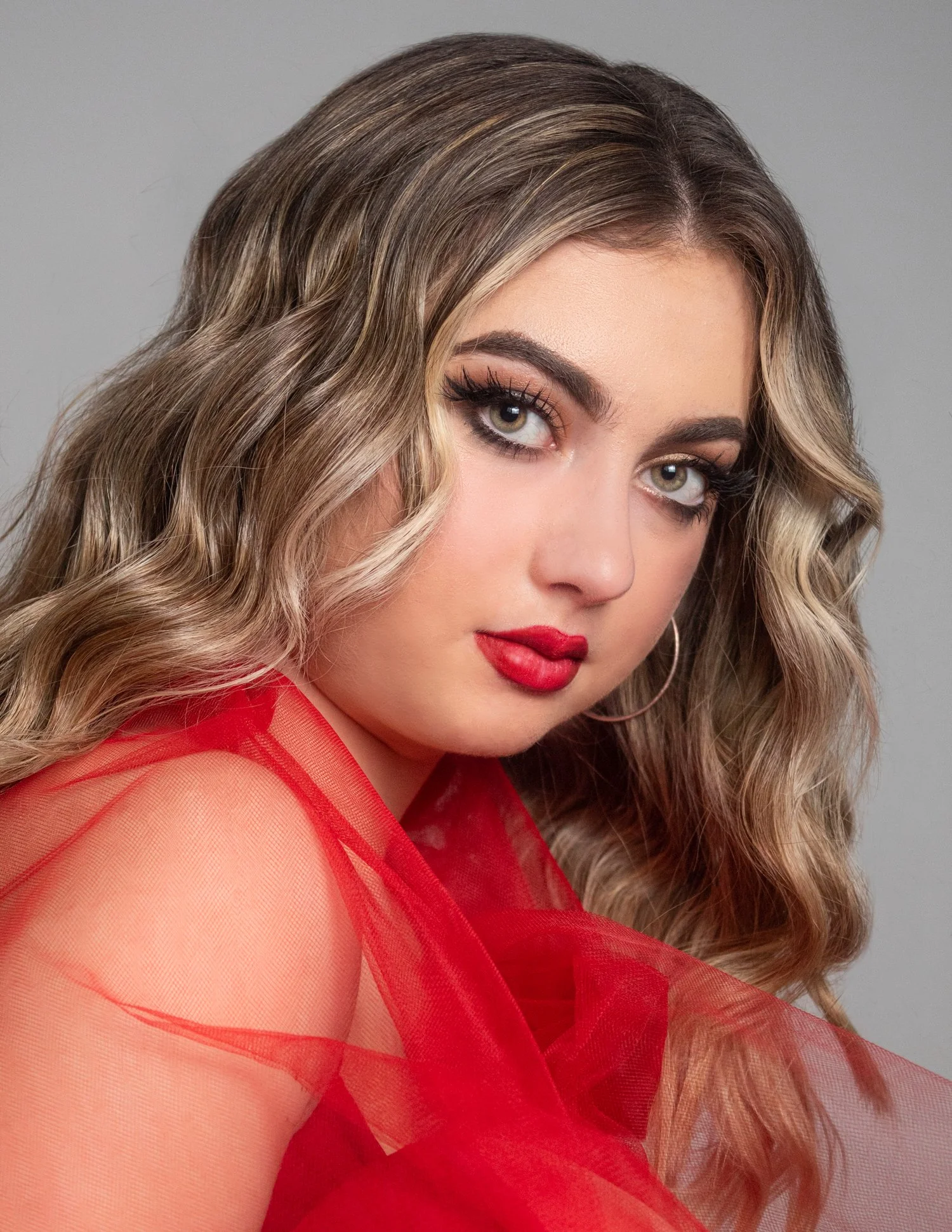 A woman with wavy light brown hair and green eyes, wearing bold red lipstick, eye makeup, and a sheer red fabric over her shoulder.