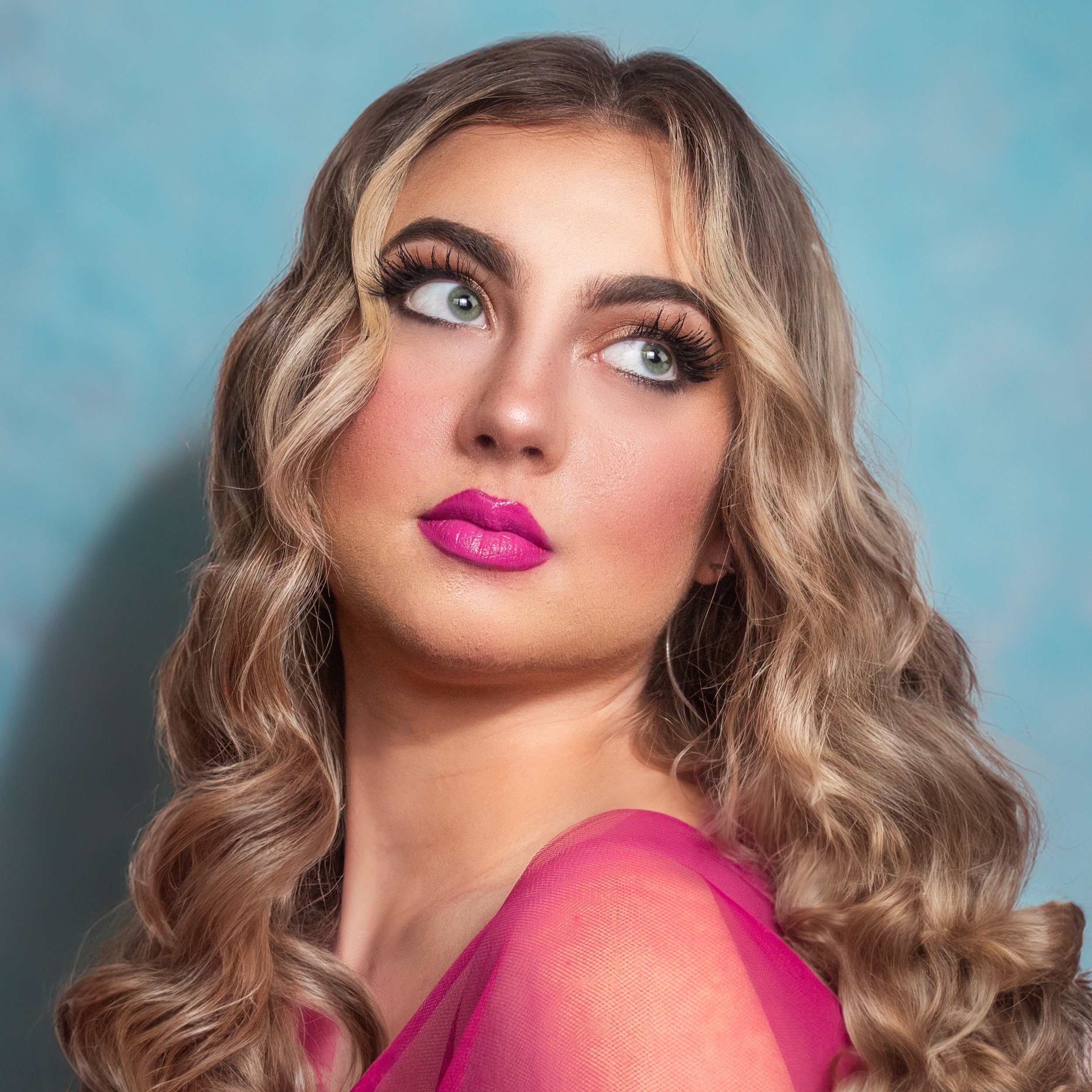 Close-up of a woman with curly blonde hair, radiant makeup including pink lipstick and dramatic eye makeup, against a blue background.