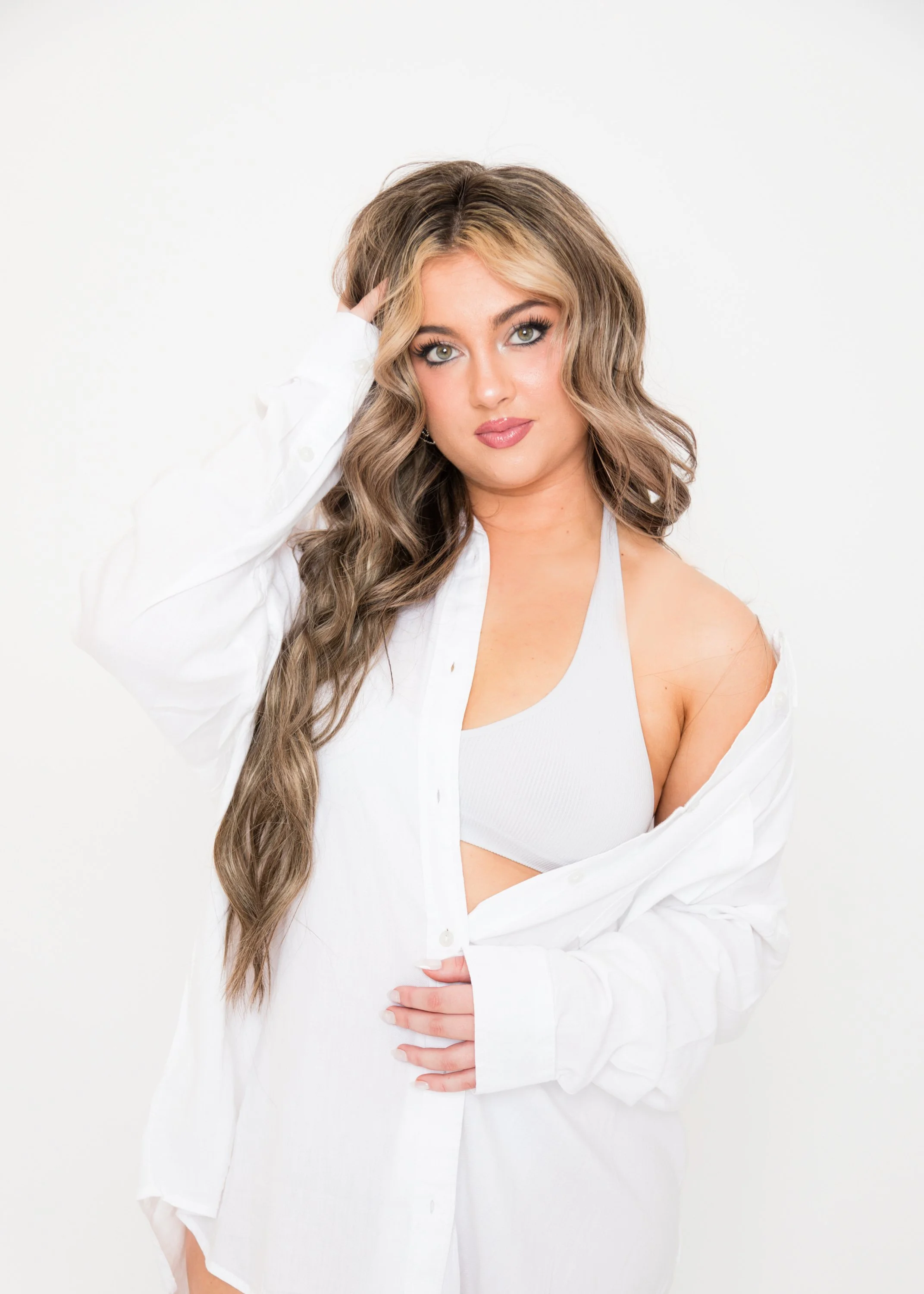 A young woman with wavy, light brown hair and blue eyes, wearing a white sports bra and a loose white button-up shirt, standing against a plain white background.