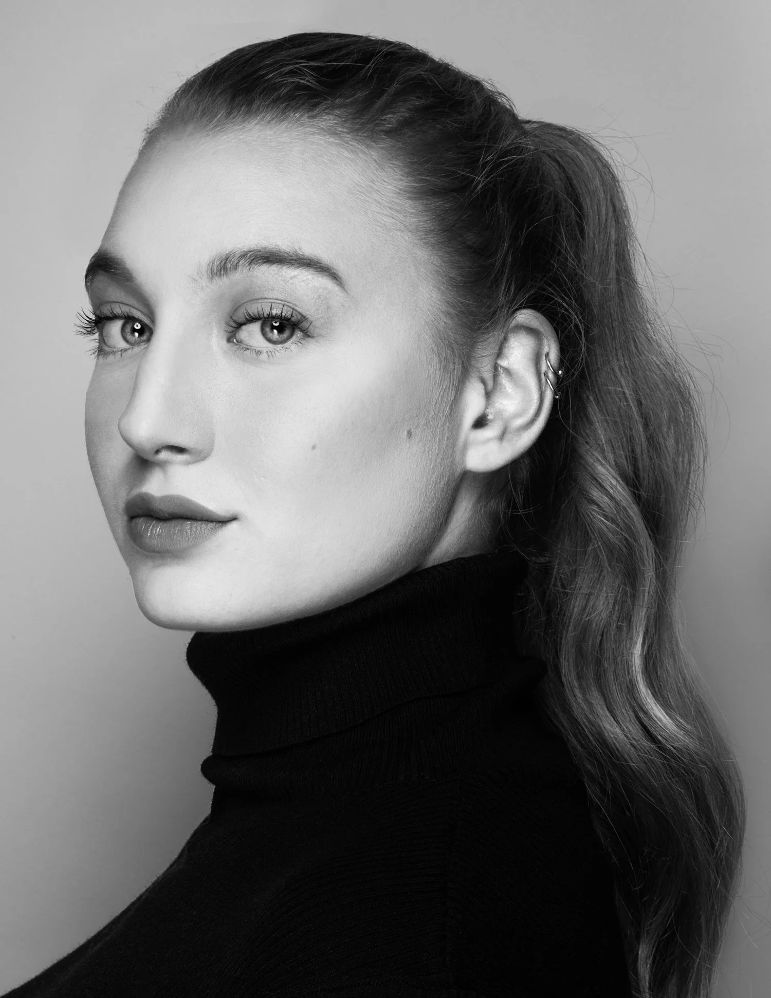 Black and white portrait of a young woman with wavy hair pulled back, wearing a turtleneck, looking at the camera.