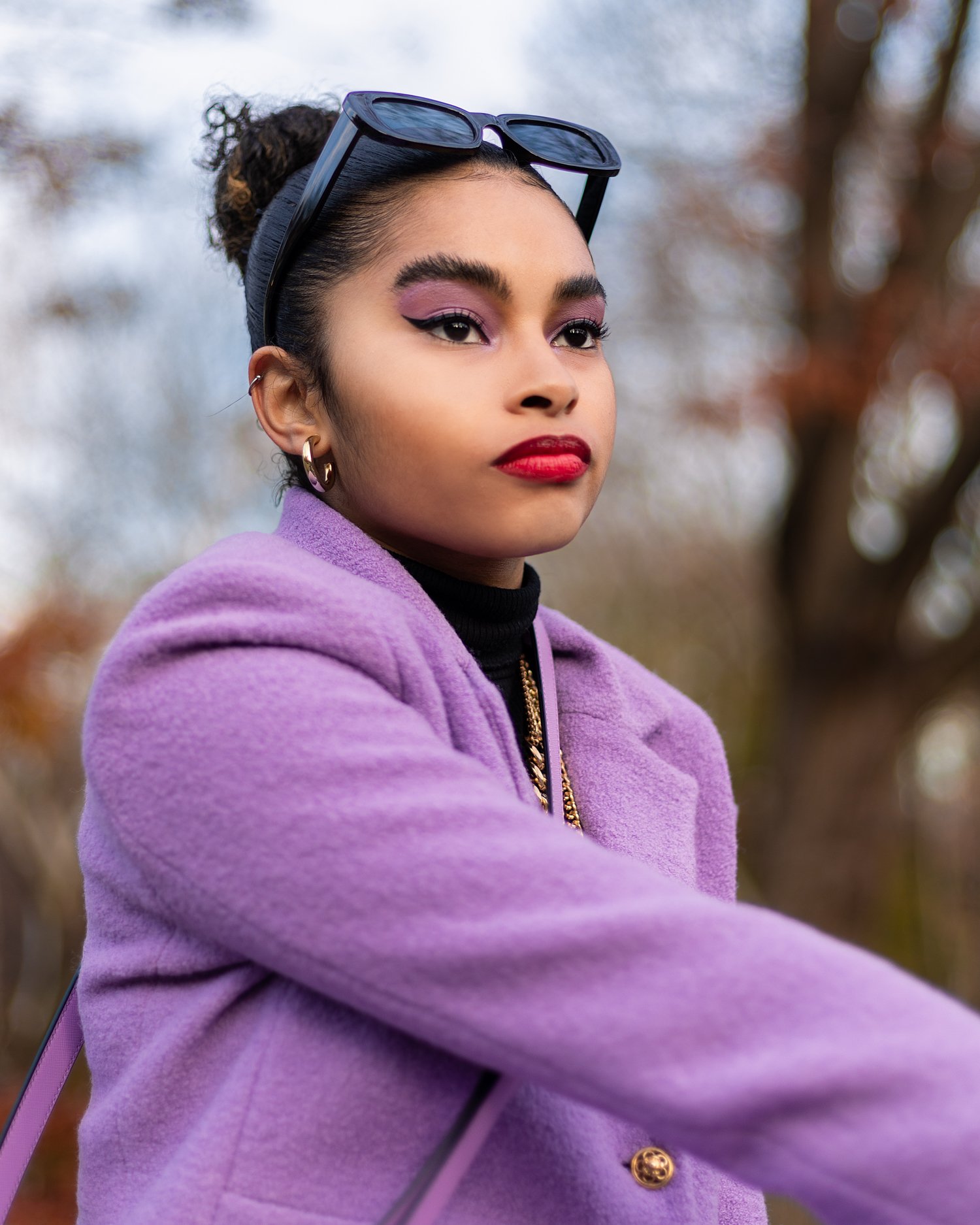 Young woman with dark curly hair in a bun, wearing sunglasses on her head, purple coat, bold makeup, and red lipstick, outdoors during daytime.