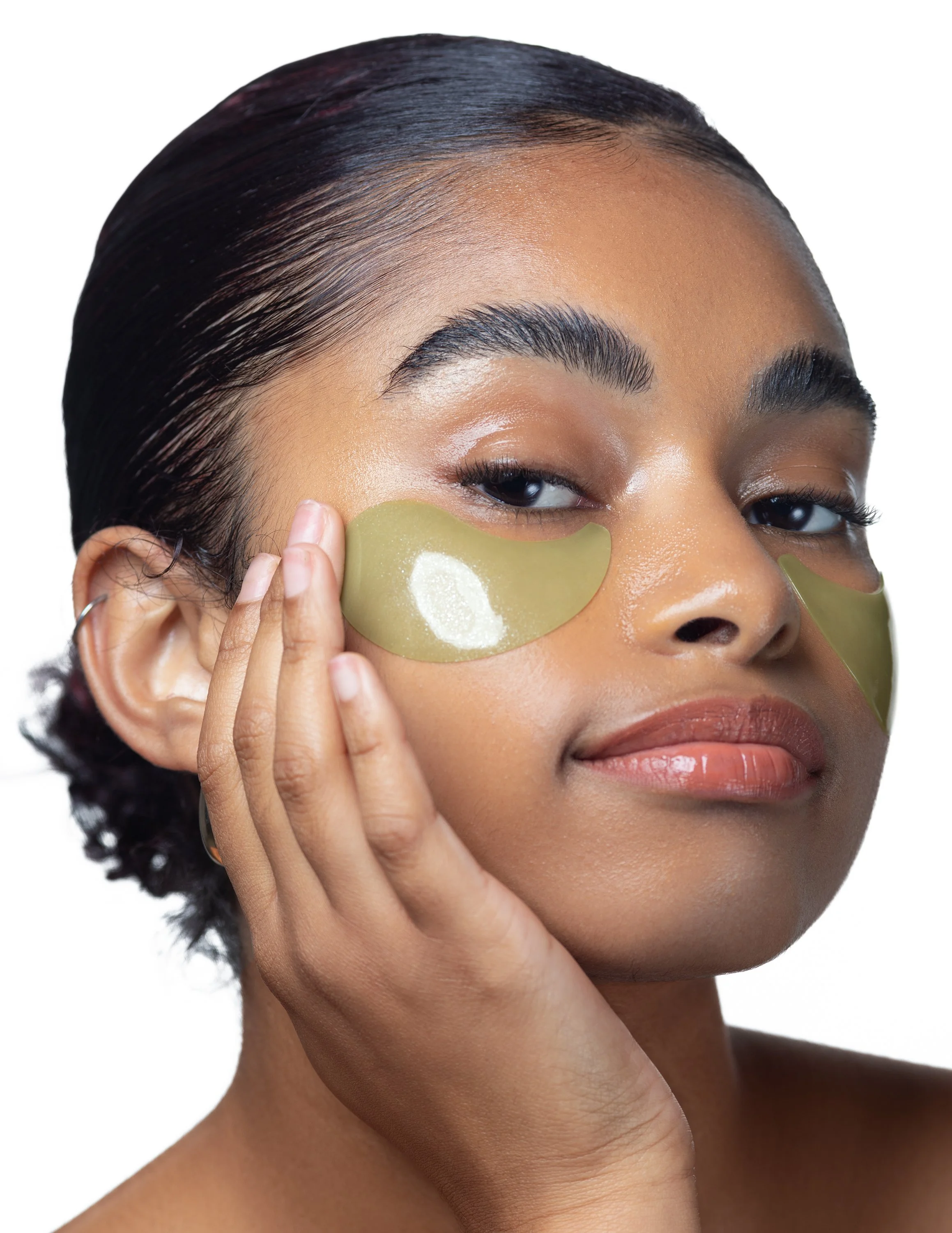 Close-up of a woman with short hair applying eye patches under her eyes, looking at the camera with a slight smile.