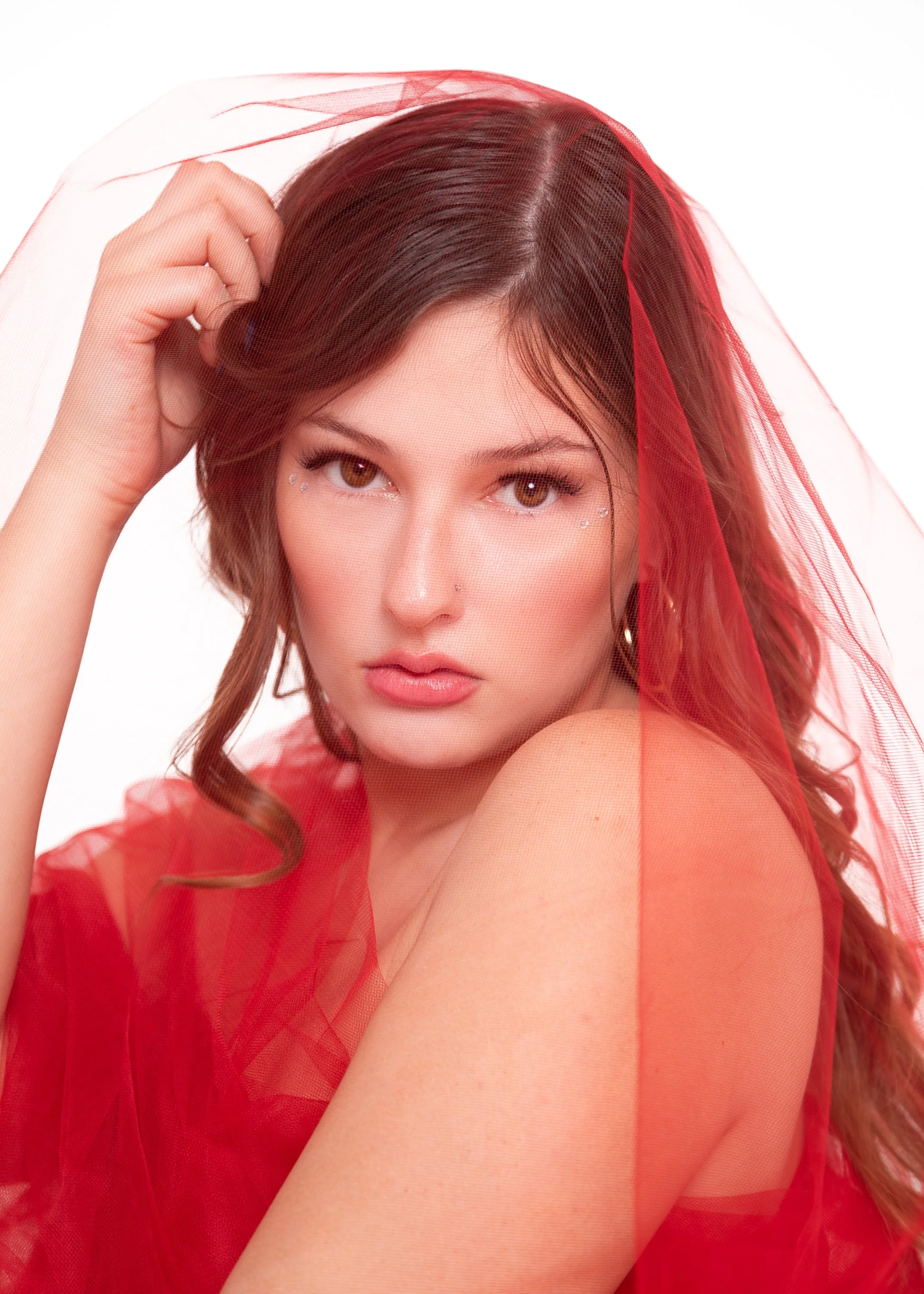 A woman with brown hair and brown eyes, wearing a red sheer veil and top, looking directly at the camera with a serious expression, against a plain white background.