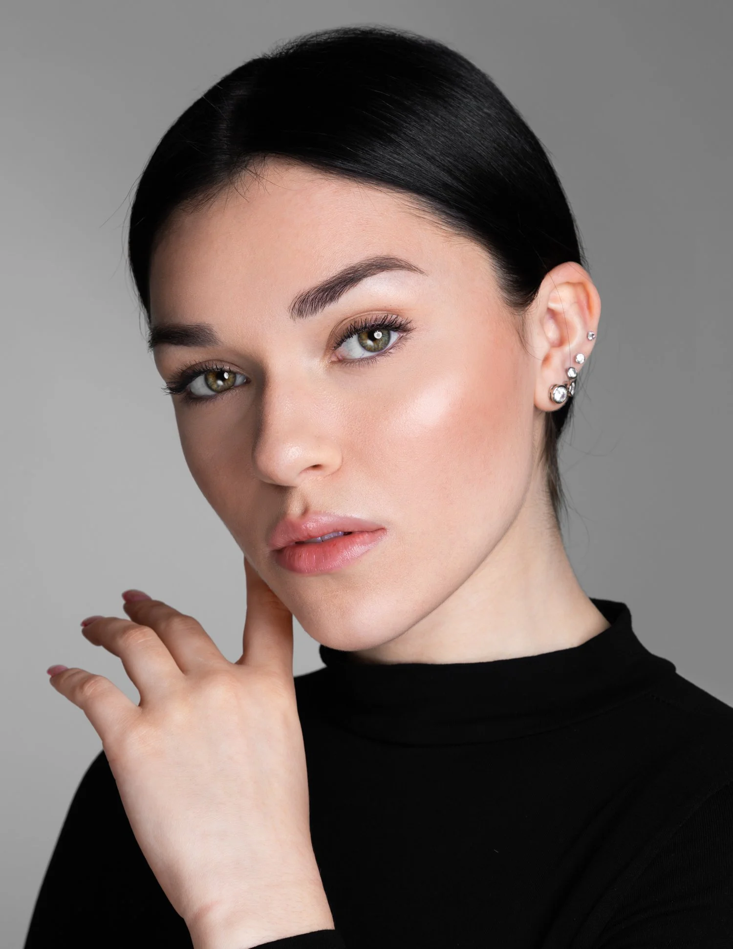 Close-up portrait of a woman with fair skin, black hair, and earrings, wearing a black turtleneck, looking directly at the camera.