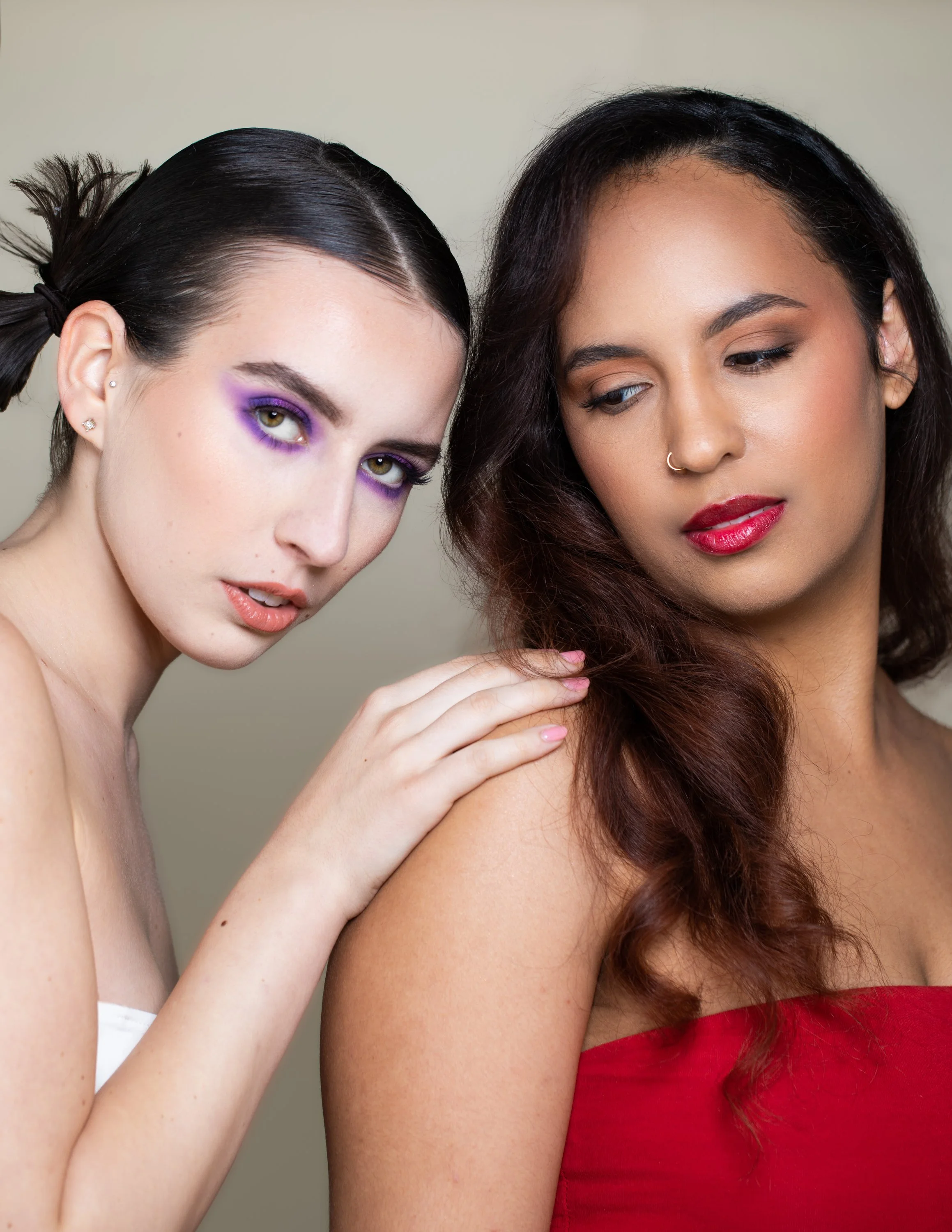 Two women with makeup, one with purple eyeshadow and the other with red lipstick, touching shoulder of the other
