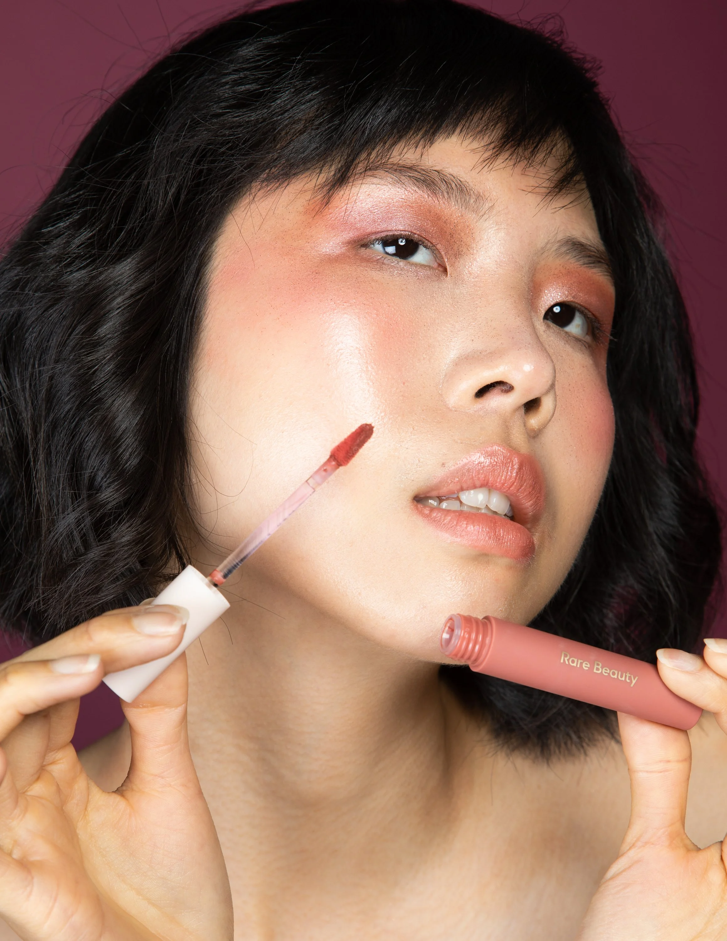 Close-up of a woman with short dark hair applying lip gloss, holding a tube labeled 'Rare Beauty,' with makeup that includes blush and eyeshadow, against a pink background.