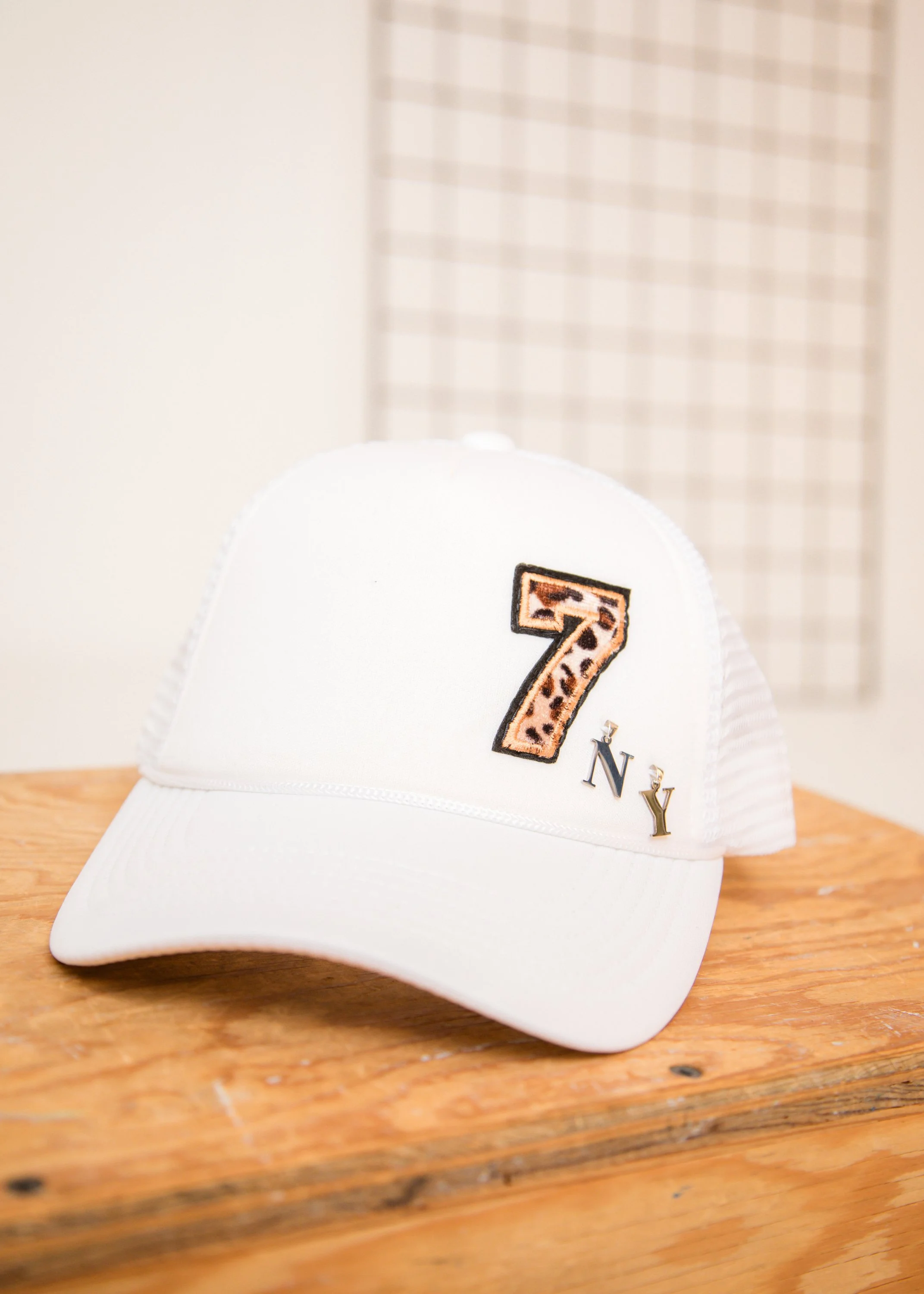 A white trucker hat with a leopard print number 7 and the word 'NY' embroidered on the front, placed on a wooden surface.