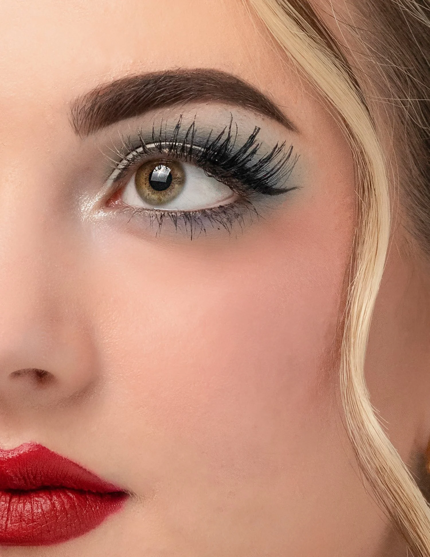 Close-up of a woman's eye with makeup, including eye shadow, eyeliner, mascara, and red lipstick, with a curled blonde hair strand beside her face.