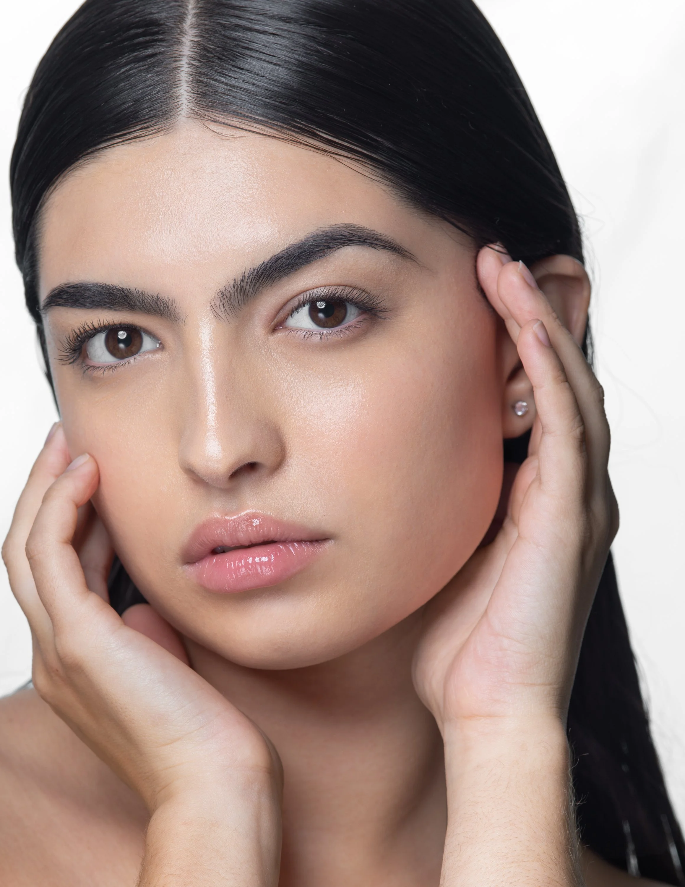 Close-up of a woman with dark hair, touching her face with both hands, showing her clear skin and natural makeup.