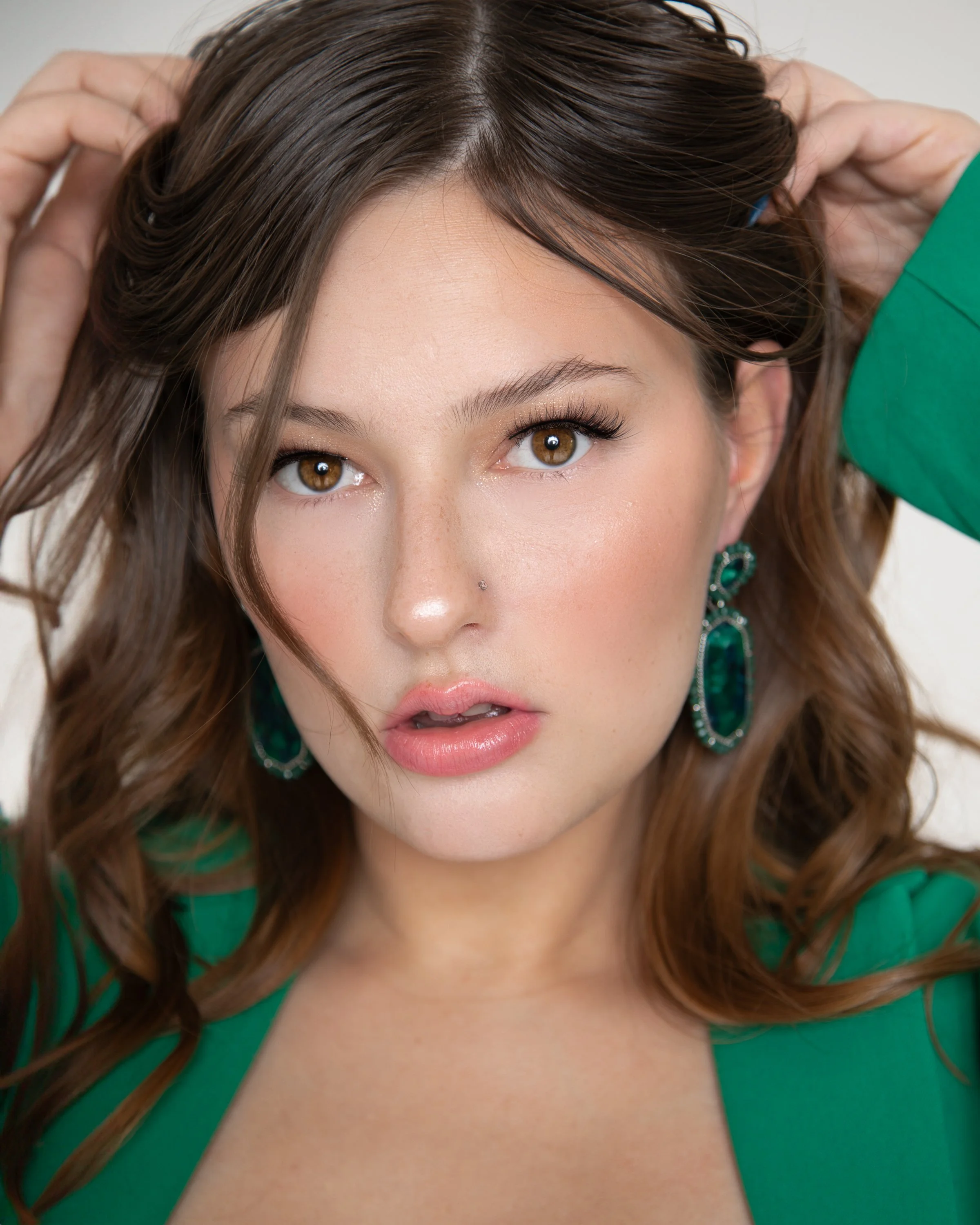 A woman with brown hair and brown eyes looking directly at the camera, touching her hair with both hands, wearing green earrings and a matching green outfit.