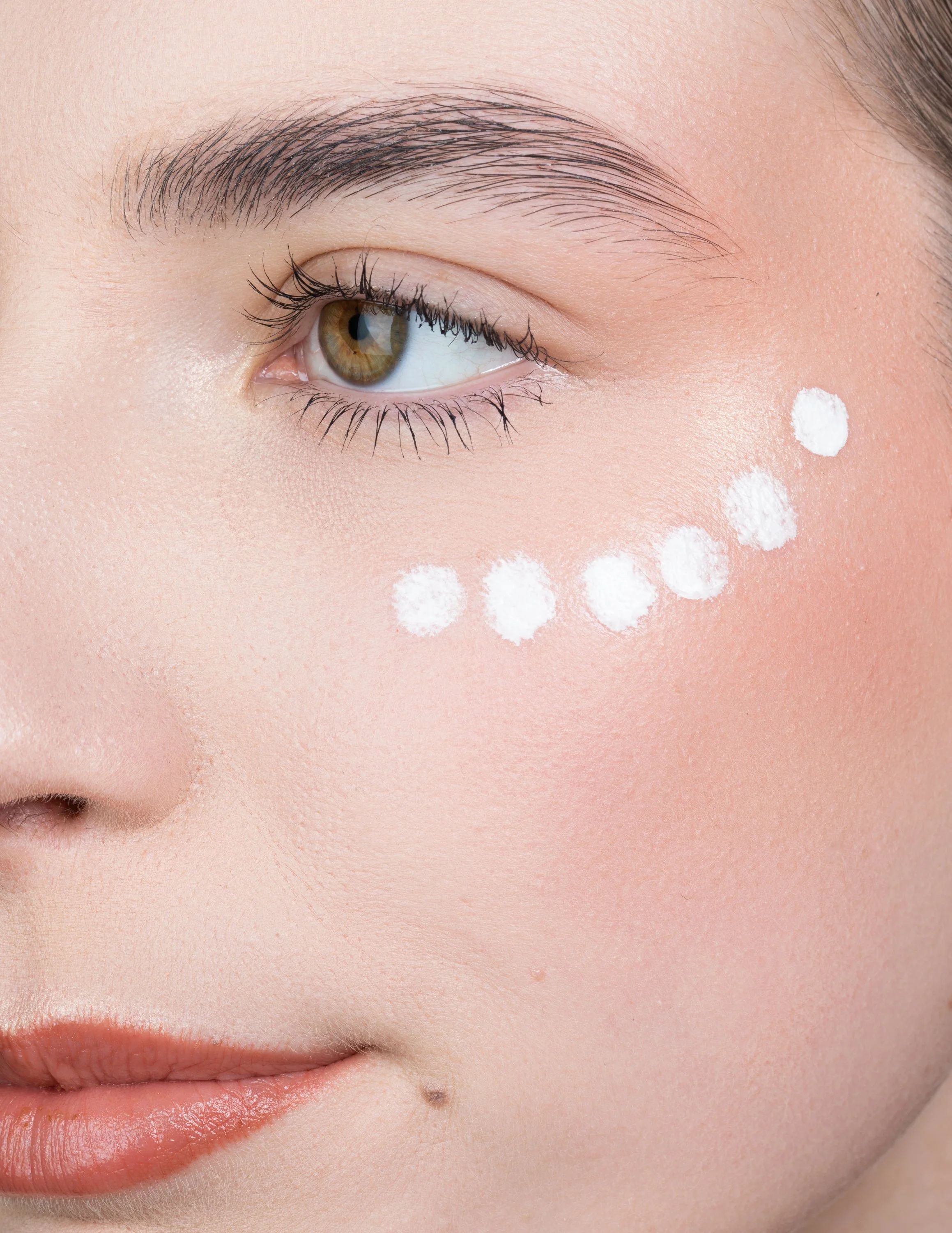 Close-up of a woman's face with white dots of cosmetic product under her eye.