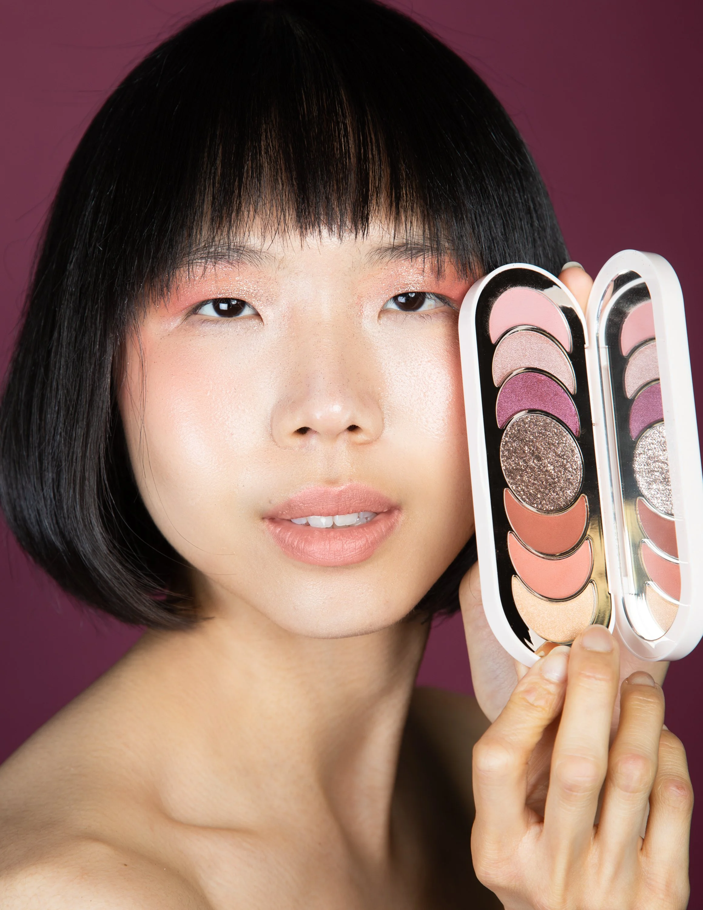 A woman with short black hair and makeup holding an open eyeshadow palette with various pink, purple, and glitter shades against a purple background.