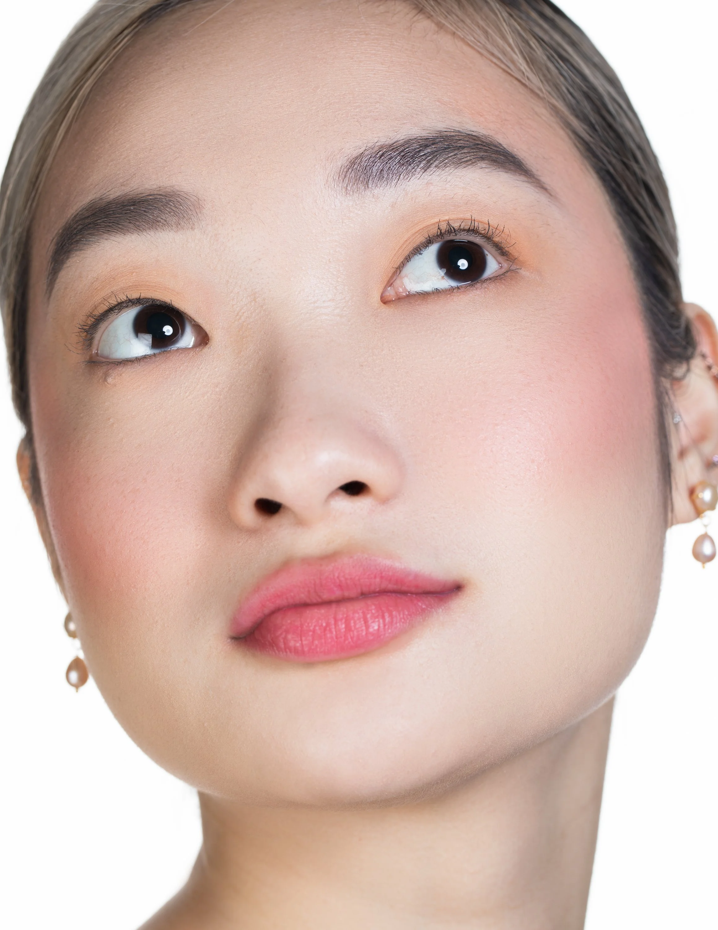Close-up of a young woman's face with clear skin, makeup, and jewelry, looking upward.