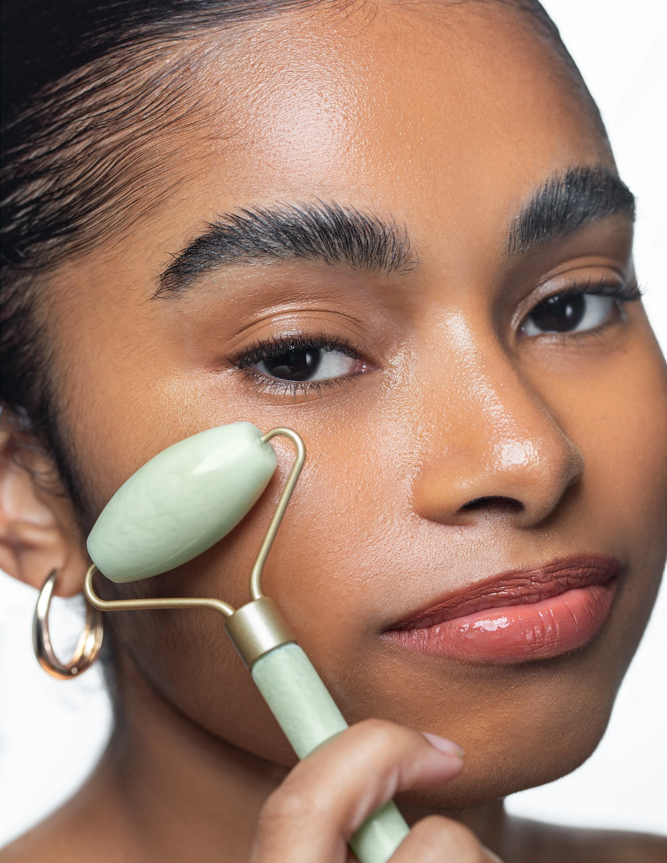 Close-up of a woman using a jade roller on her face, with smooth skin, well-groomed eyebrows, and a neutral expression.