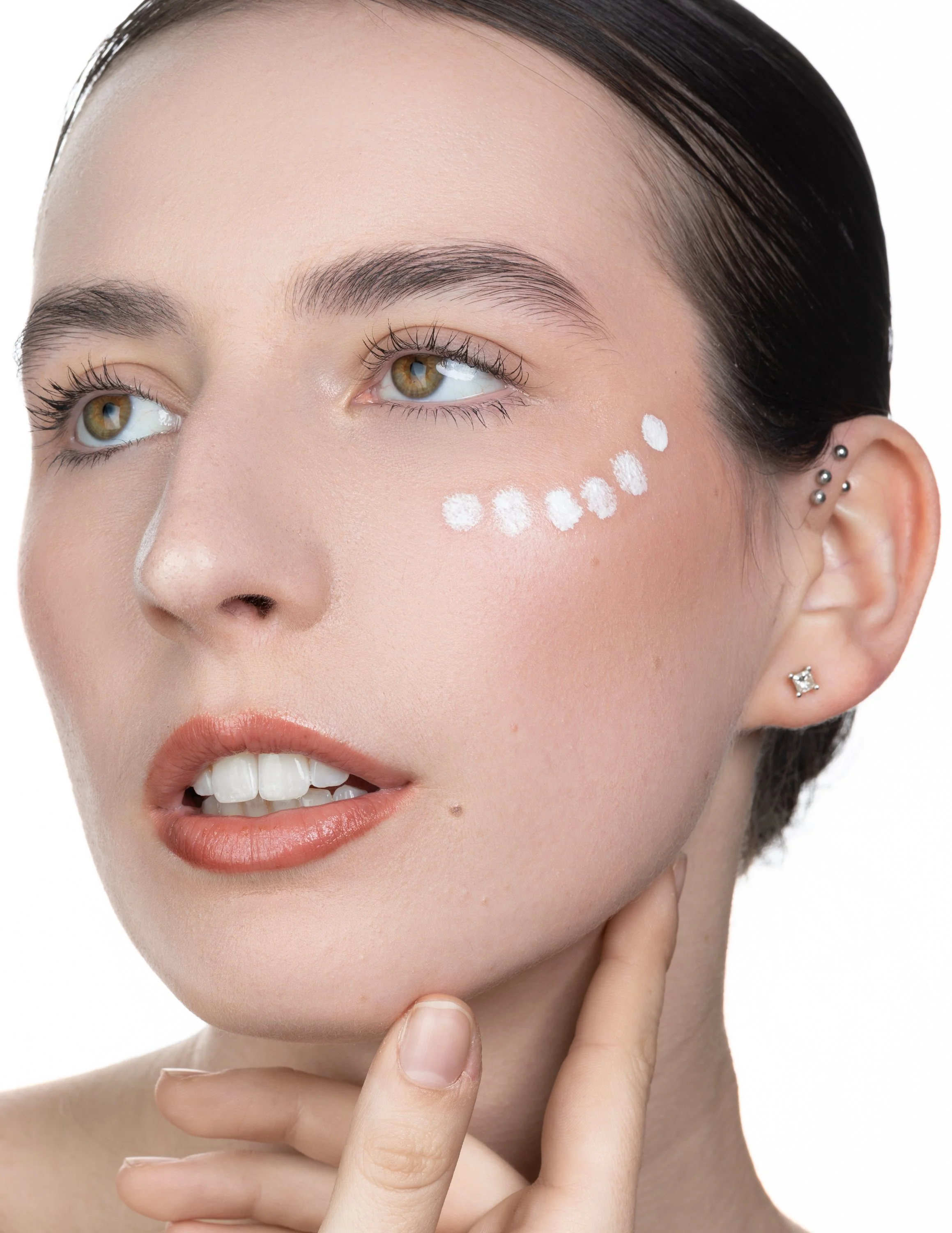 A woman with brown hair, light makeup, and multiple earrings touching her chin. She has white dots painted on her cheek, likely for makeup or skincare purposes.