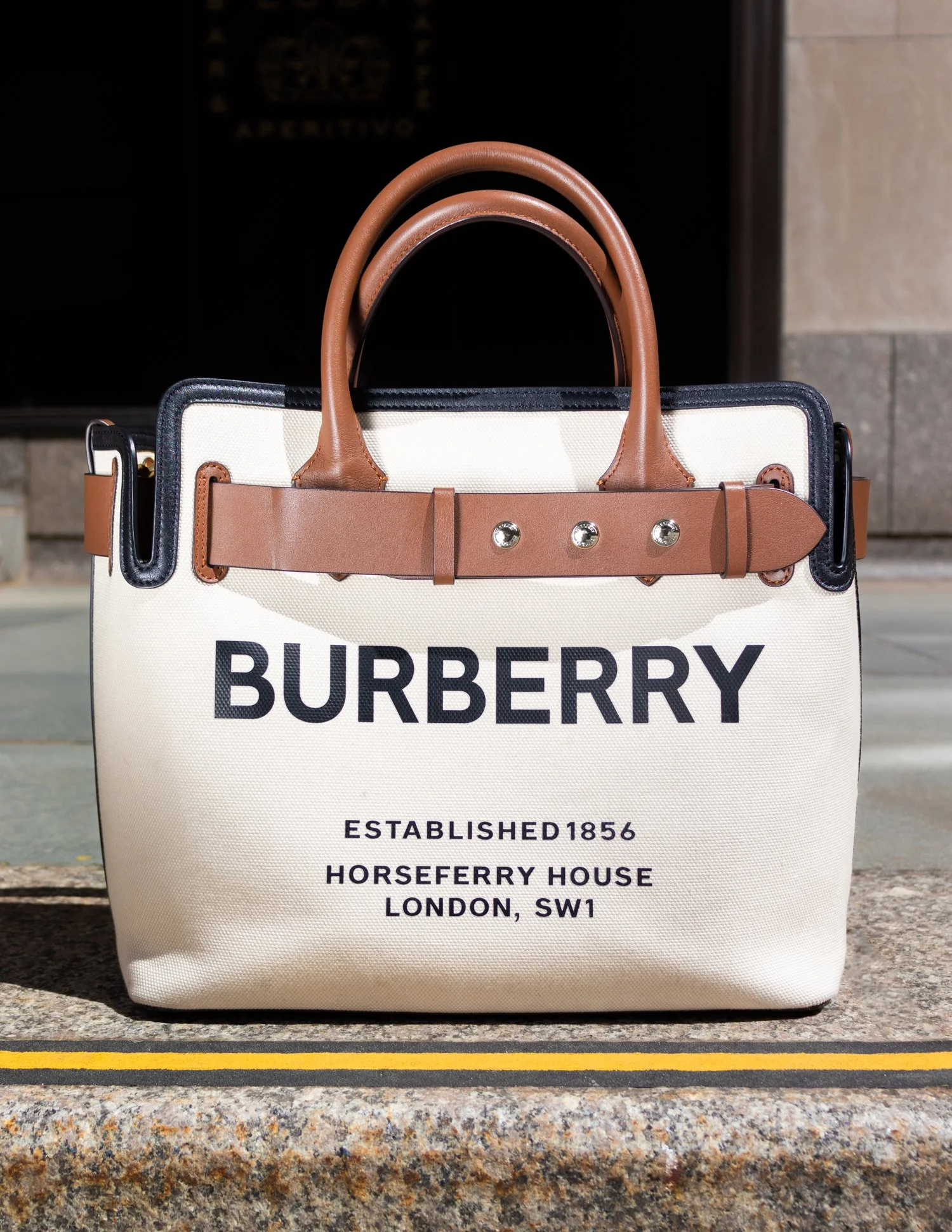 A Burberry tote bag with tan leather handles and black trim, featuring printed text about the brand's history, placed outdoors on a concrete surface.