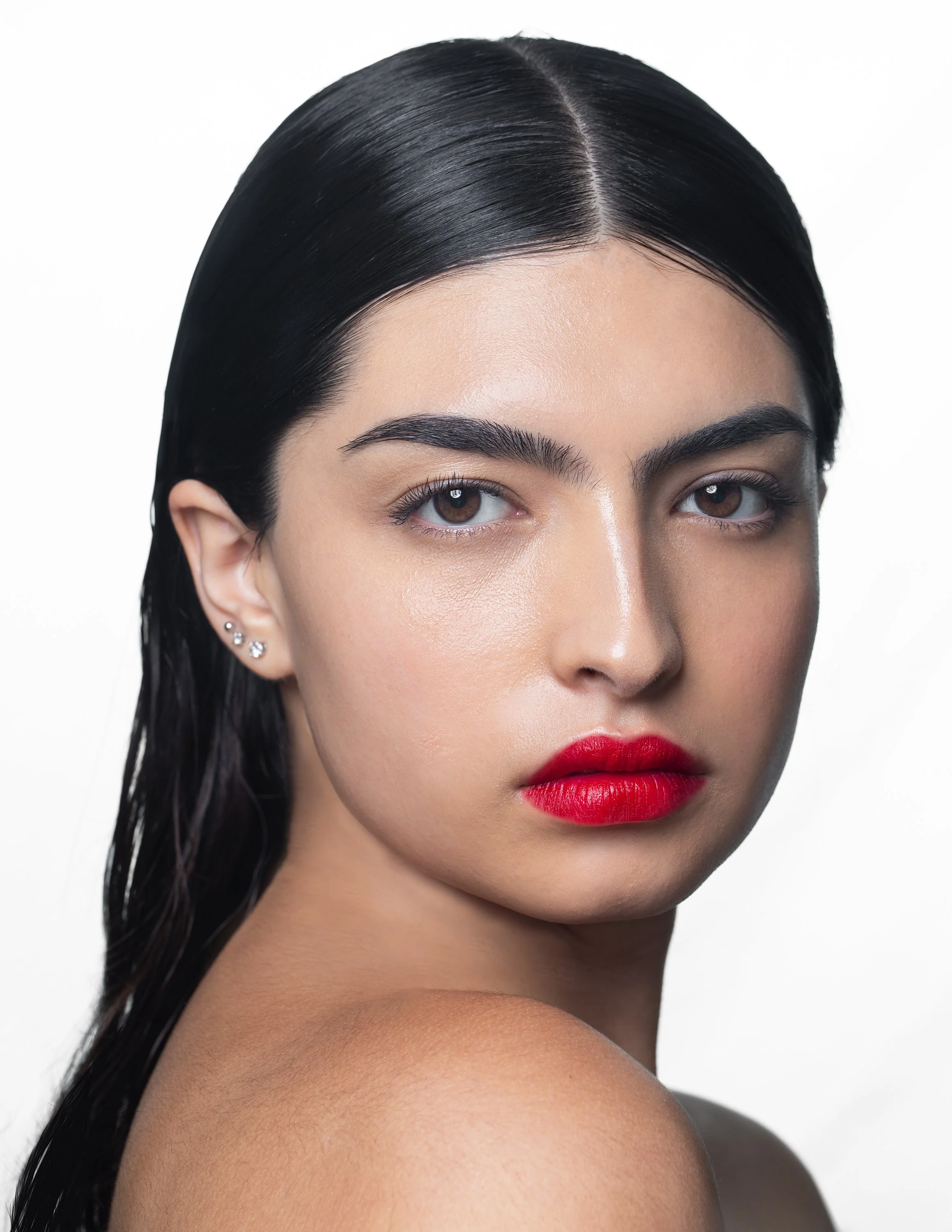 Close-up portrait of a woman with wet black hair, brown eyes, bold red lipstick, and earrings, against a white background.