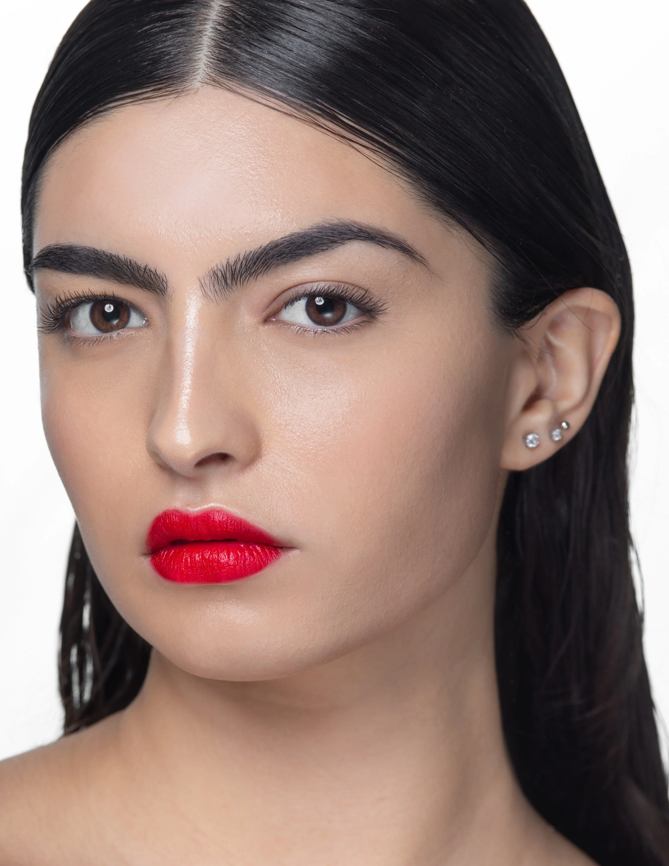 Close-up of a woman with dark brown hair, wearing red lipstick, and small earrings, against a white background.