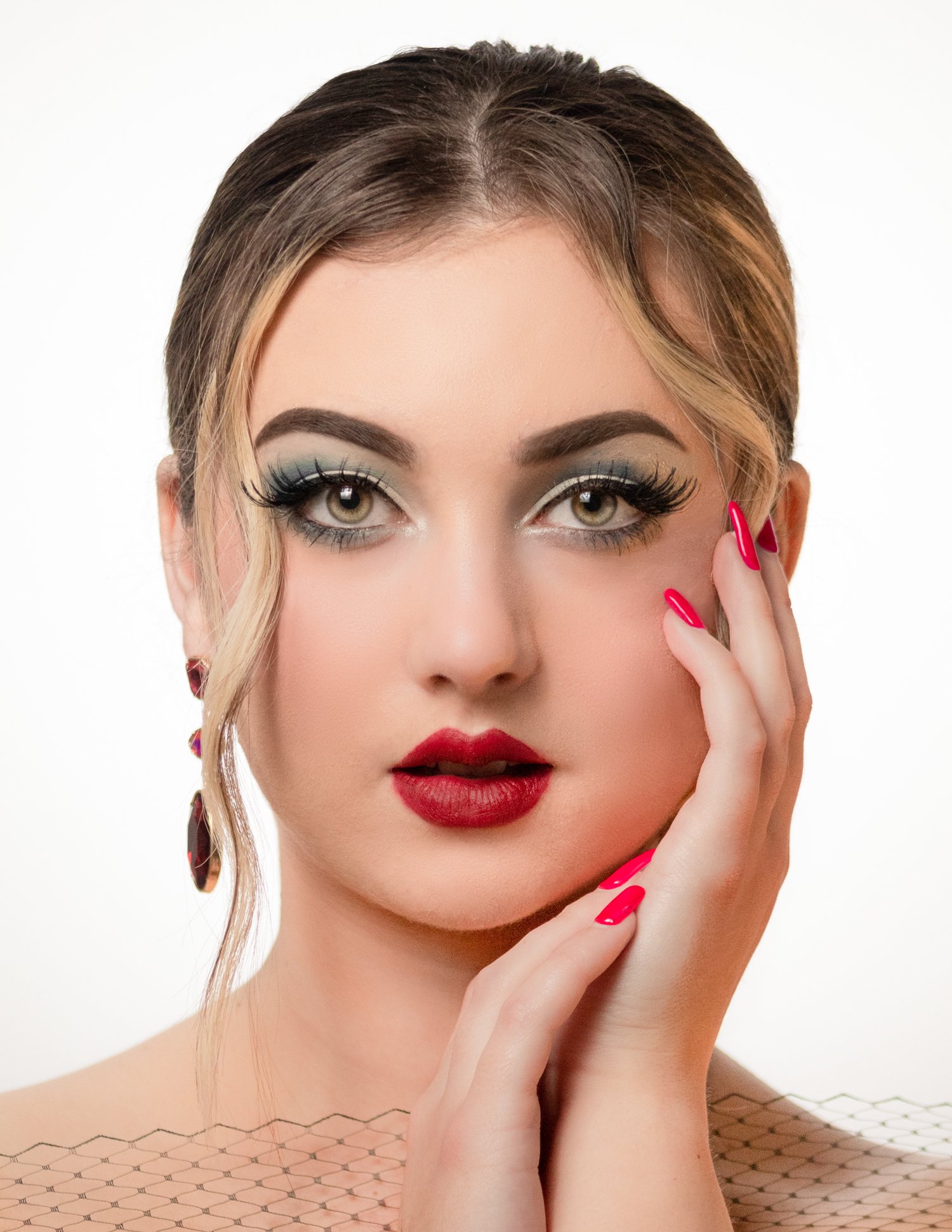 Close-up portrait of a woman with makeup, red lipstick, and long eyelashes, touching her face with her right hand, with pink manicured nails and wearing earrings, against a white background.