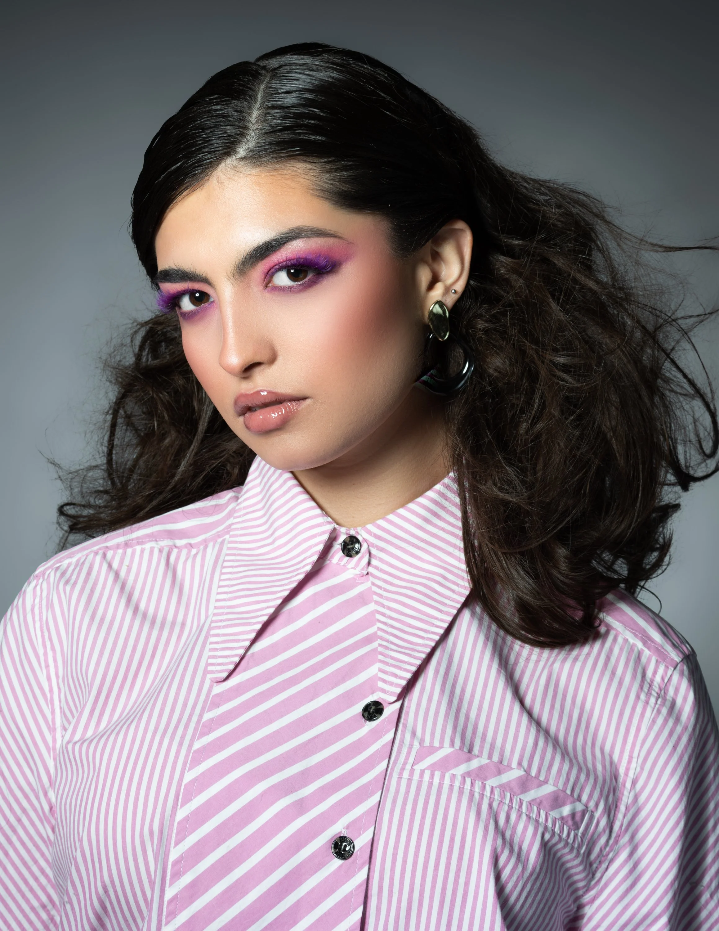 A woman with dark, wavy hair wearing pink and white striped shirt, bold purple eyeshadow, and black and green earrings, posed against a neutral gray background.