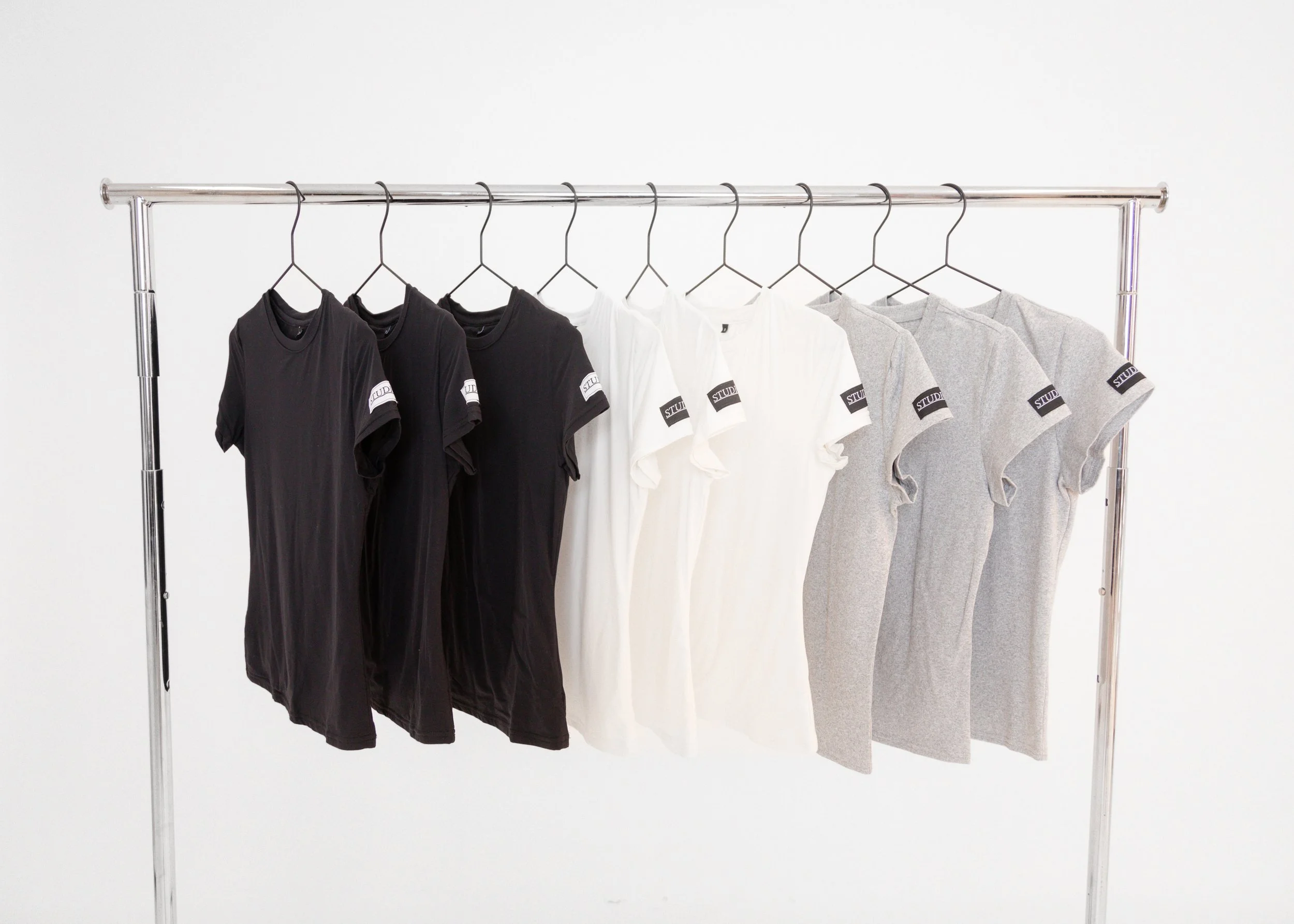 Six t-shirts hung on a metal clothing rack, with three black shirts on the left, two white shirts in the middle, and two gray shirts on the right, all with a small label on the sleeve.