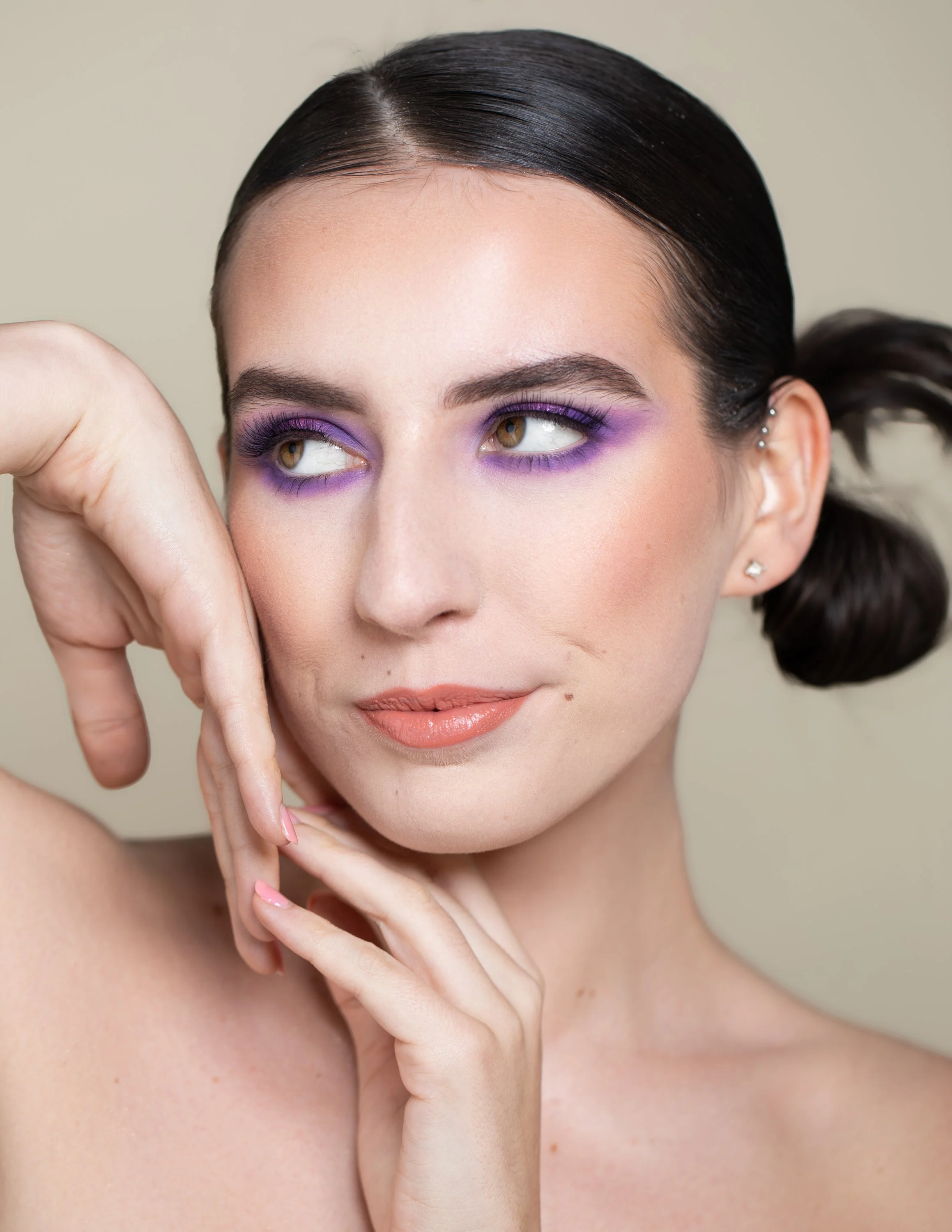 Close-up of a woman with dark hair styled in a low ponytail, wearing purple eyeshadow and nude lipstick, resting her face on her hand with fingers near her chin and cheek.