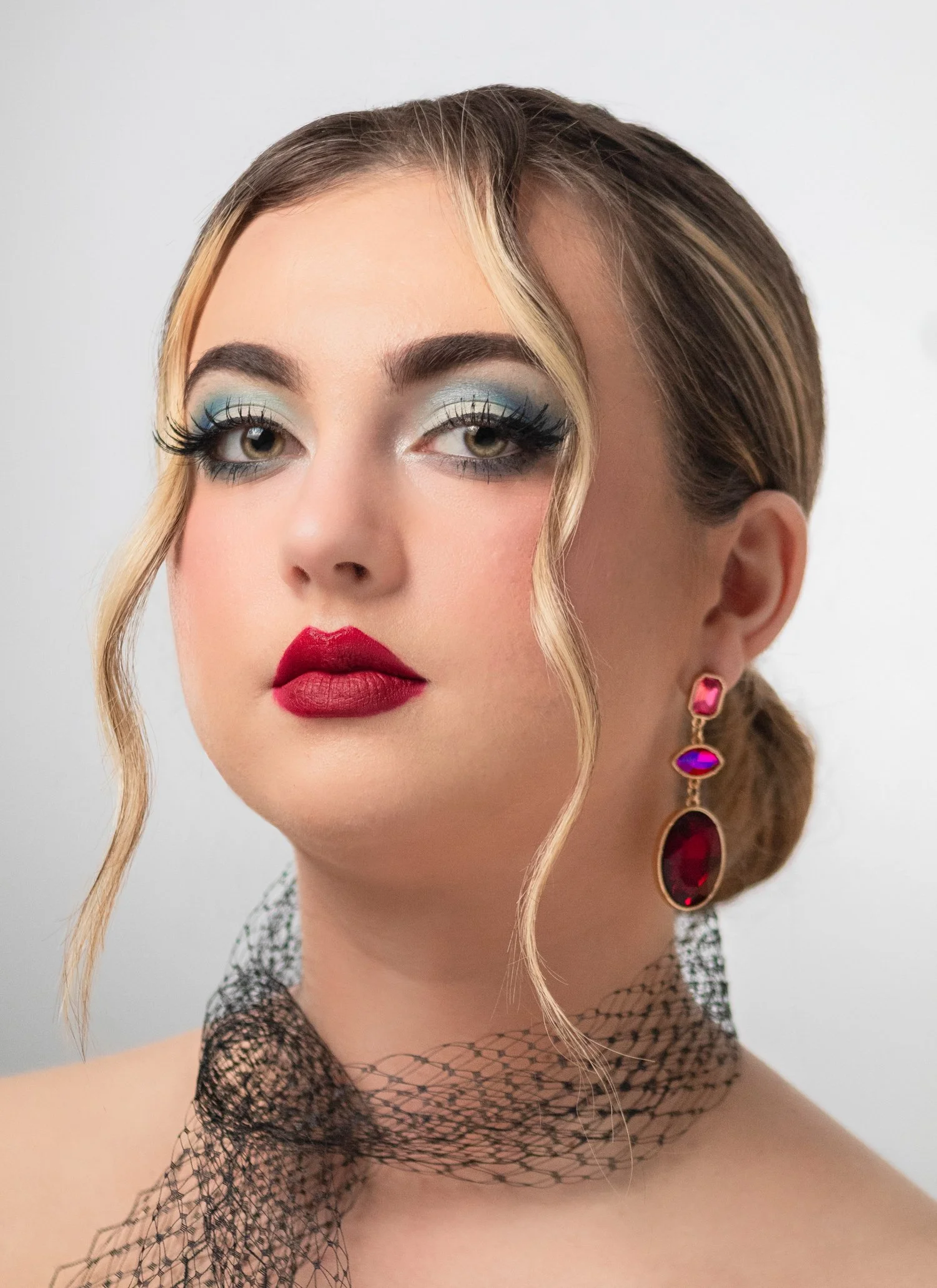 Close-up portrait of a woman with glamorous makeup, large earrings, and a black netted scarf around her neck.