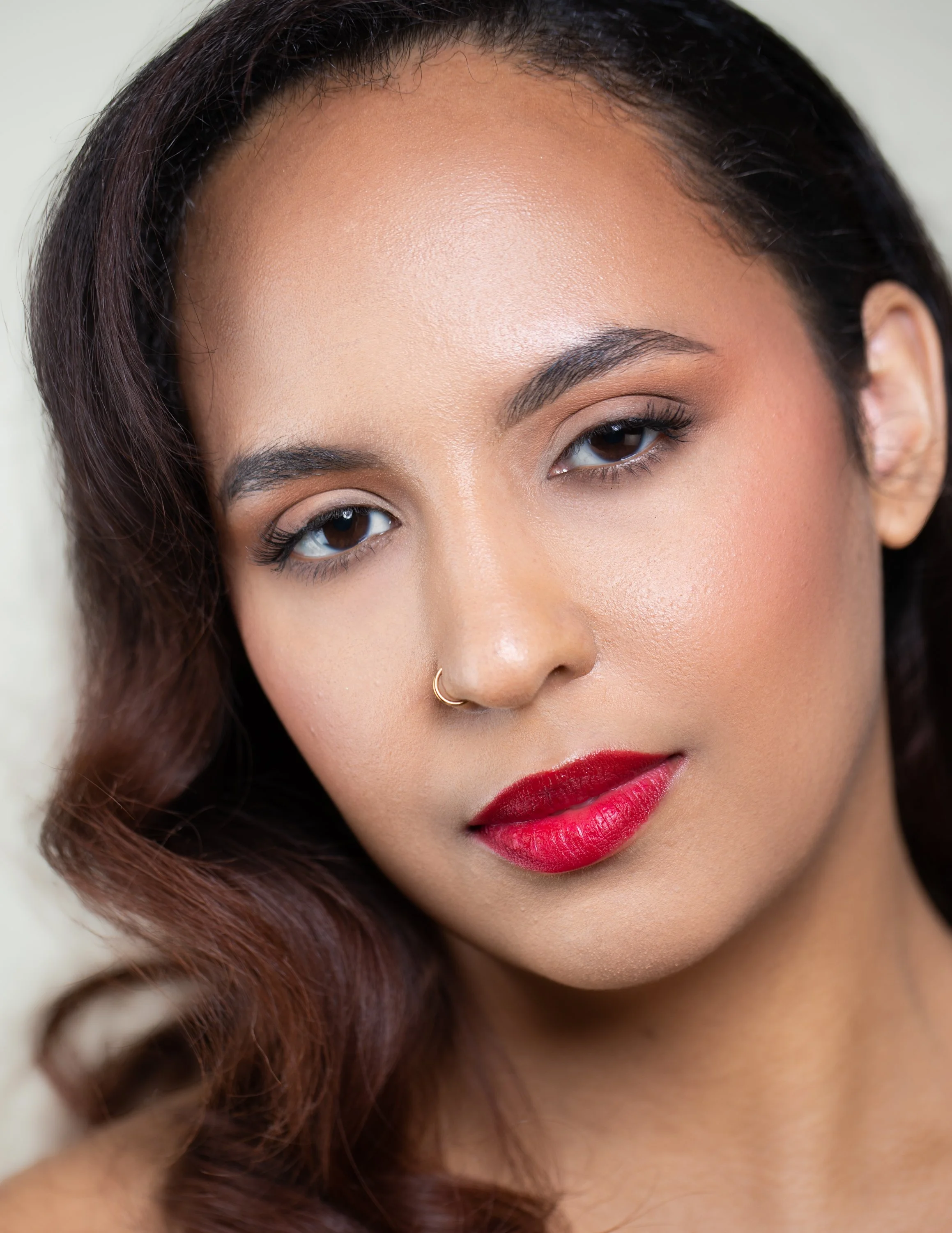 Close-up portrait of a woman with wavy brown hair, wearing makeup with red lipstick, and a small gold nose ring.