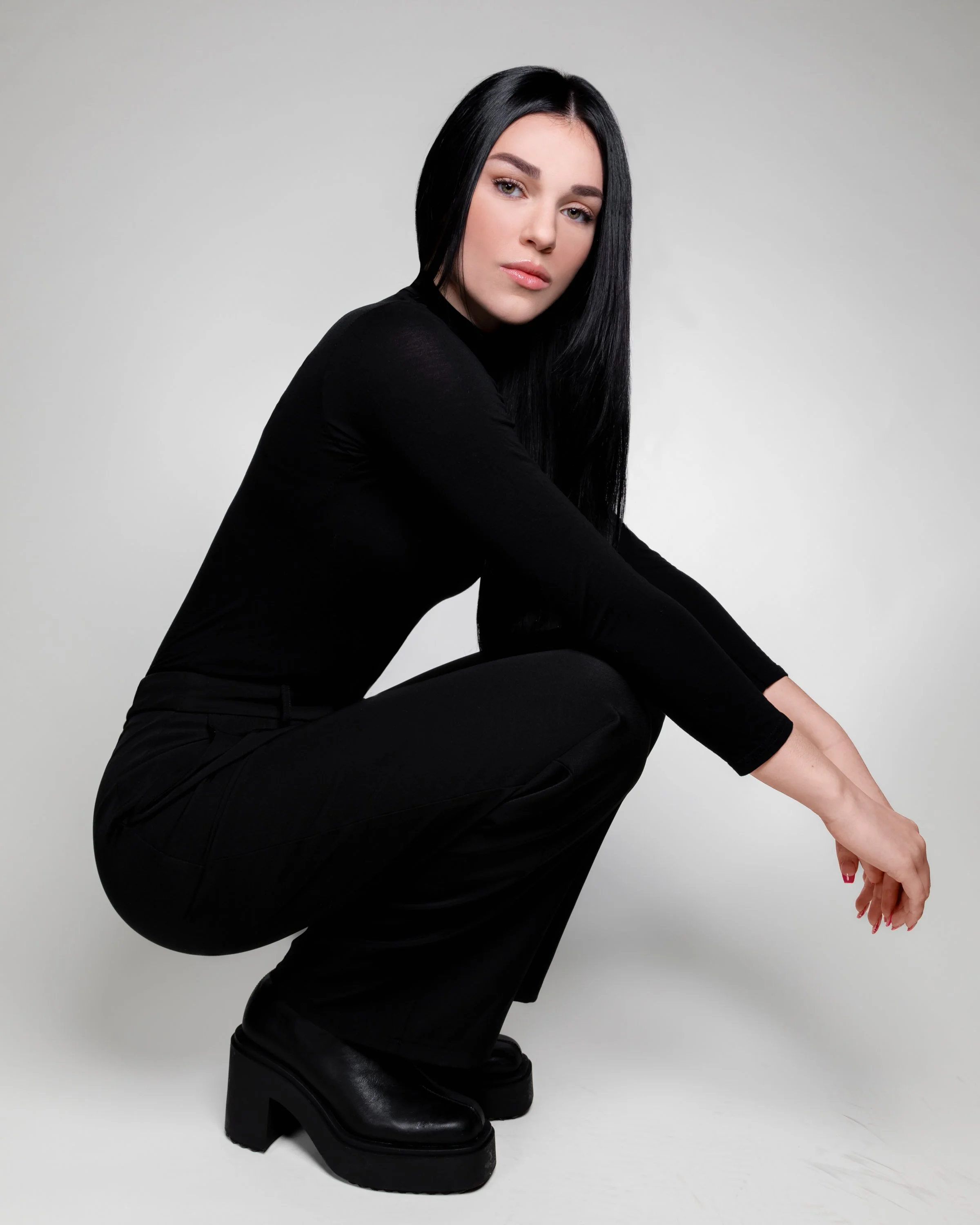 A woman with long black hair wearing a black long-sleeve top, black pants, and black platform shoes crouches and looks at the camera against a plain light gray background.