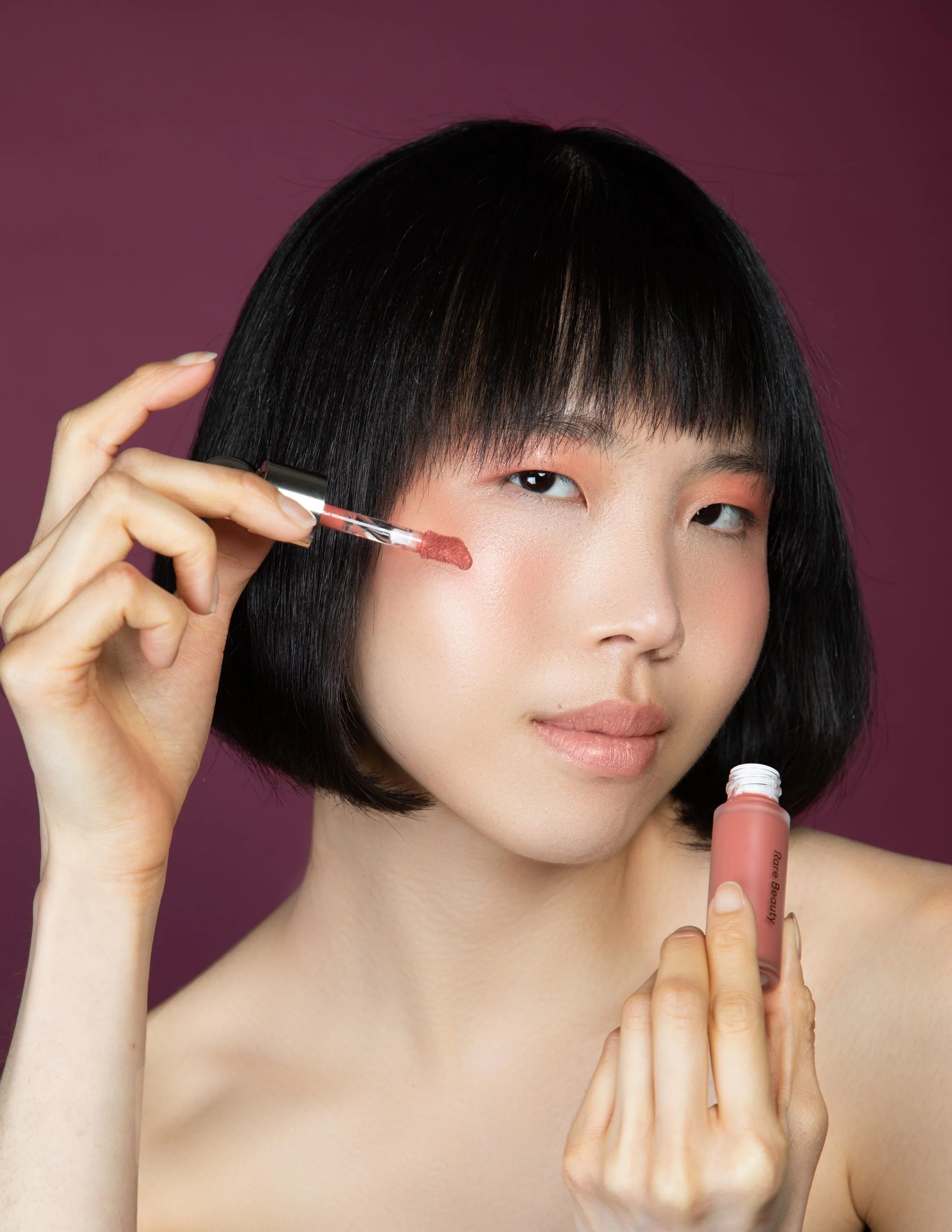 Woman with black hair applying lip gloss against a mauve background.