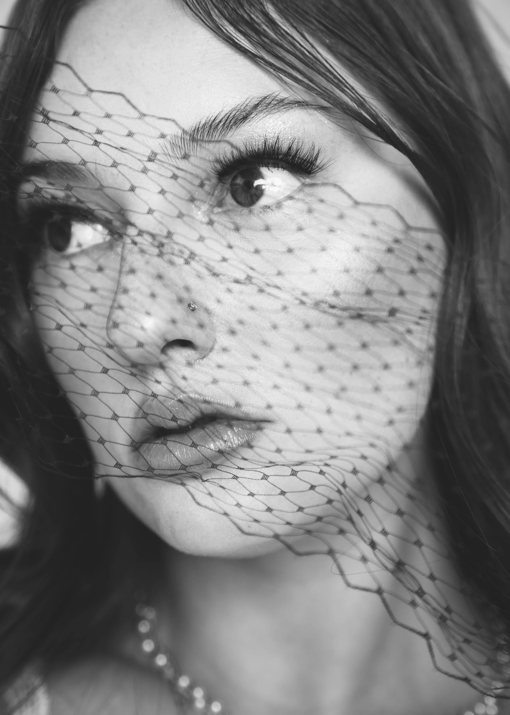 Black and white close-up portrait of a woman with a mesh veil over her face, highlighting her eye, lips, and nose piercing.