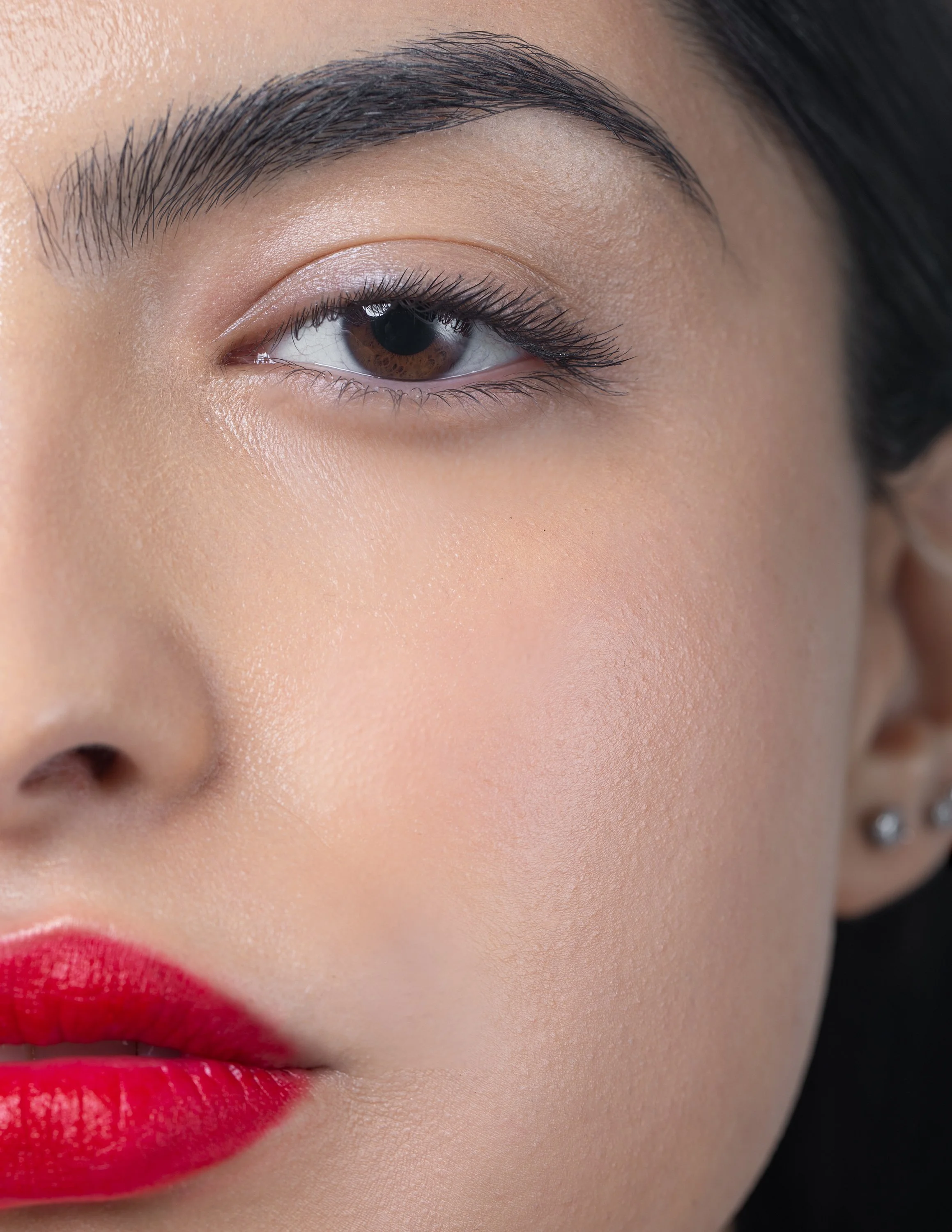 Close-up of a woman's face, focusing on her right eye, eyebrow, and lips, with smooth skin, red lipstick, and a several piercings on her ear.